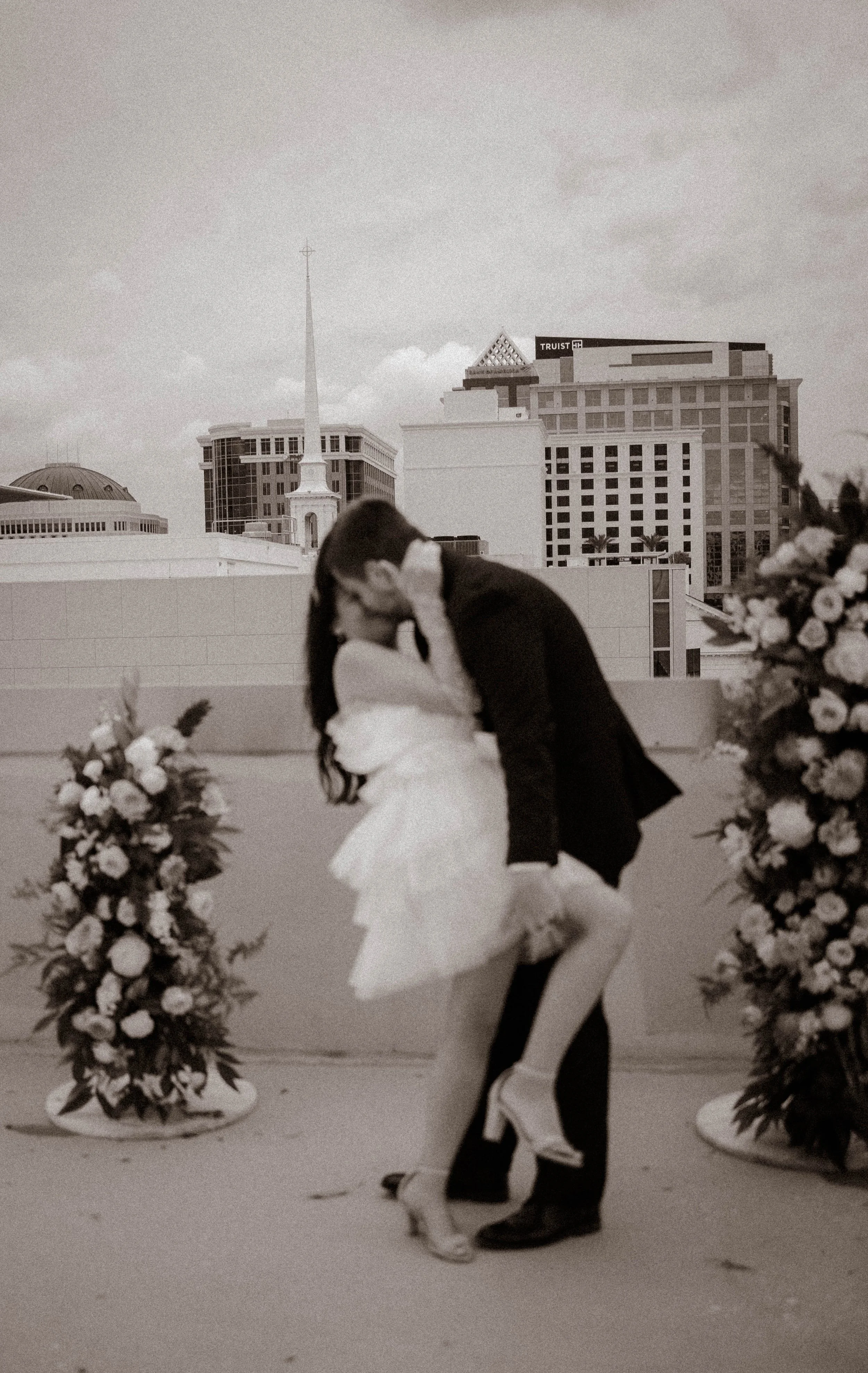 A couple dancing passionately on a rooftop, surrounded by floral arrangements, with city buildings in the background.