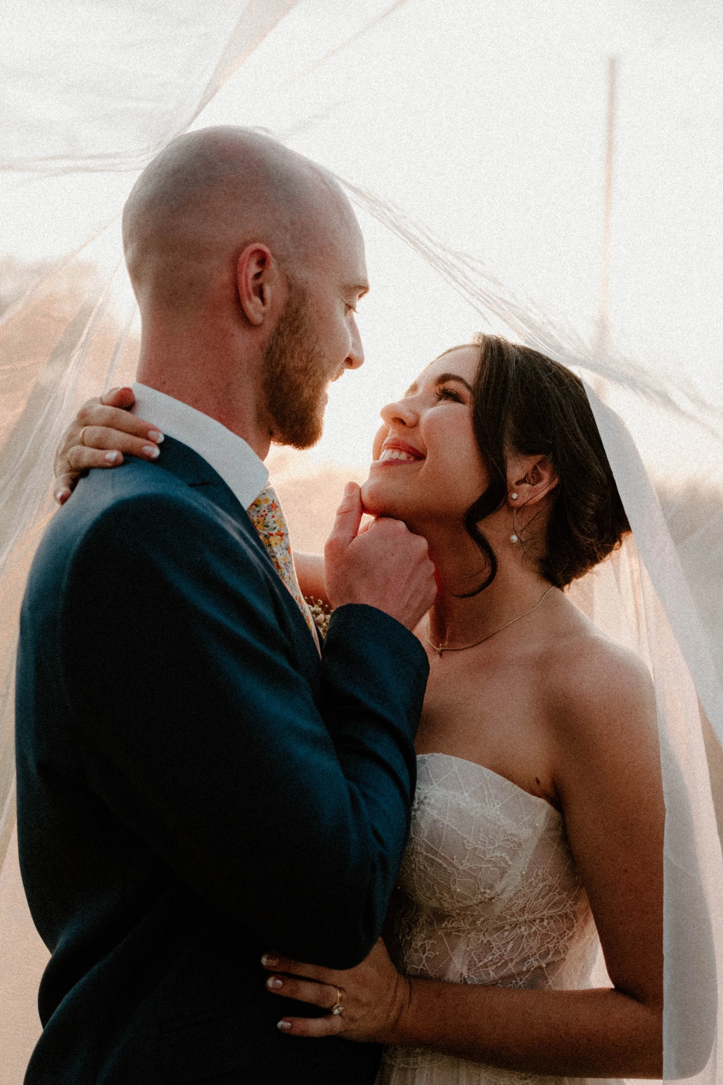 A bride and groom sharing a romantic moment under a veil, with the groom gently holding the bride's chin.
