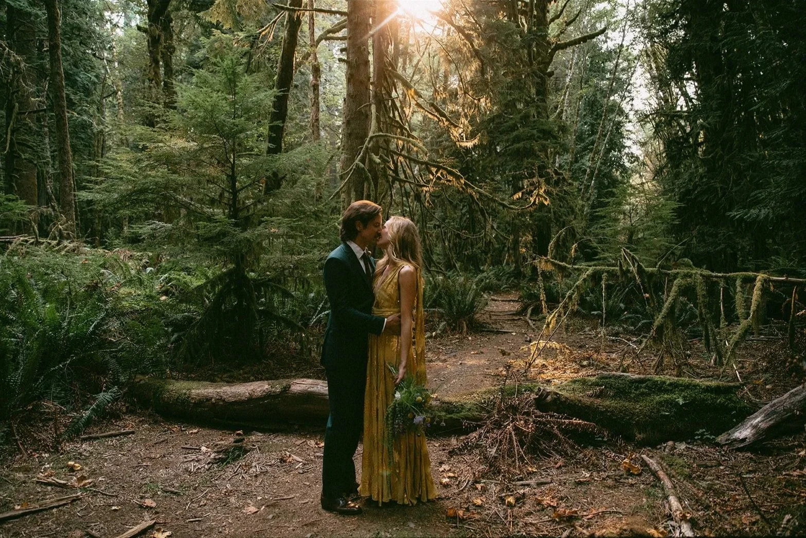 A bride in a gold dress and a groom in a green suit kissing in a forest