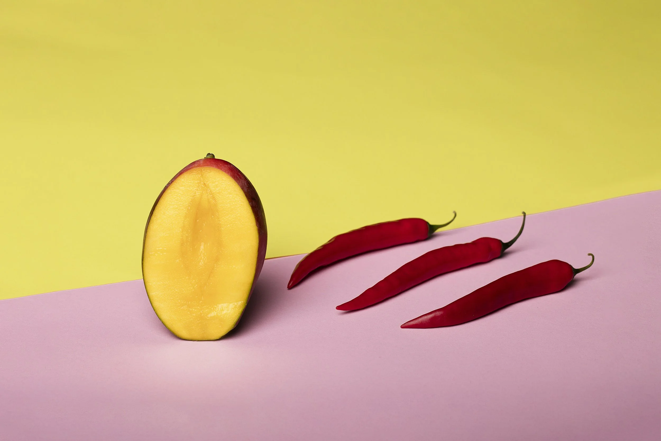 A halved yellow peach with red skin and three red chili peppers on a pink and yellow background.