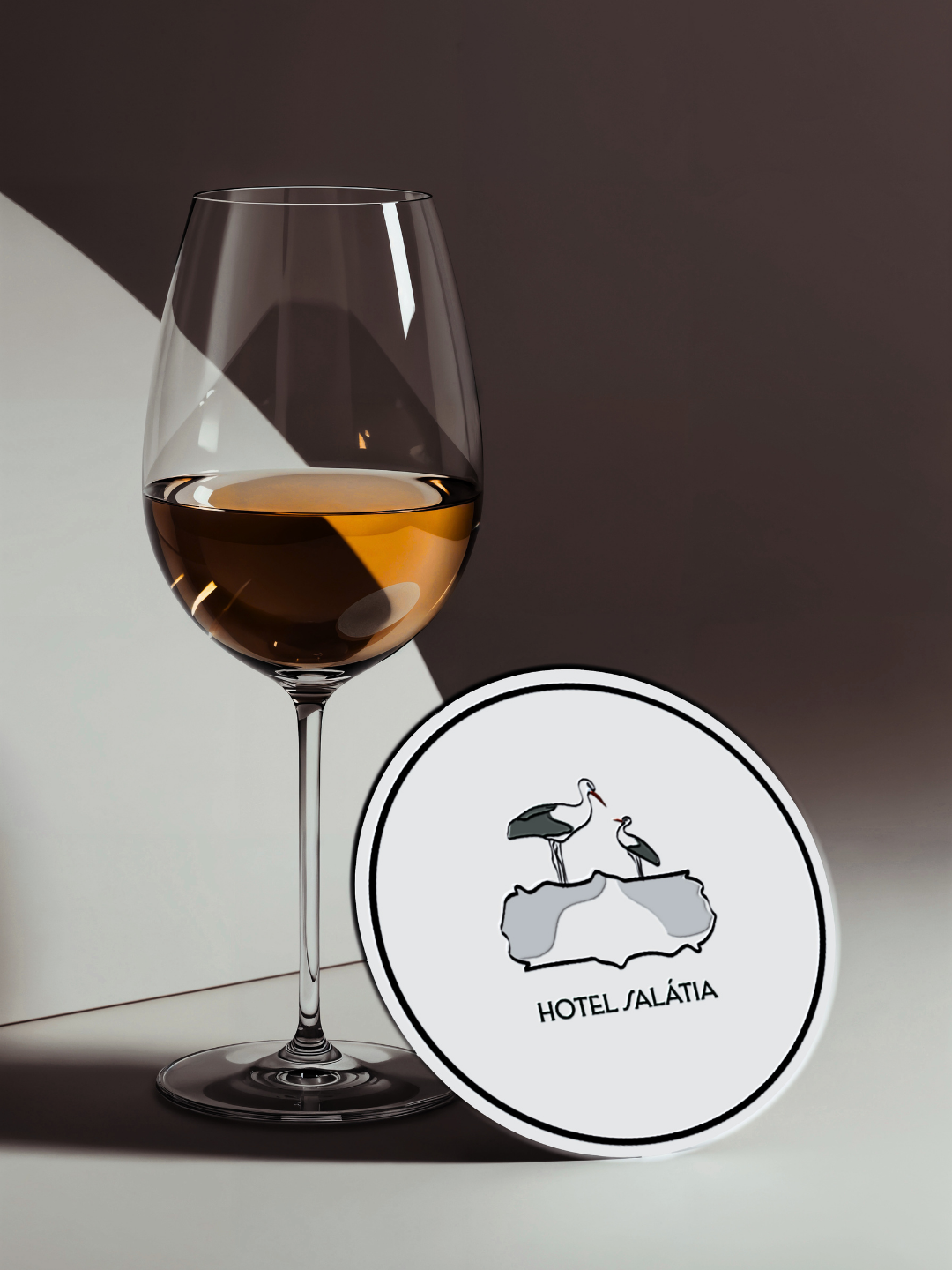 Coaster Design- Branding for Hotel Salátia. A glass of white wine on a table with a Hotel Salátia logo sticker featuring two storks on a rock.