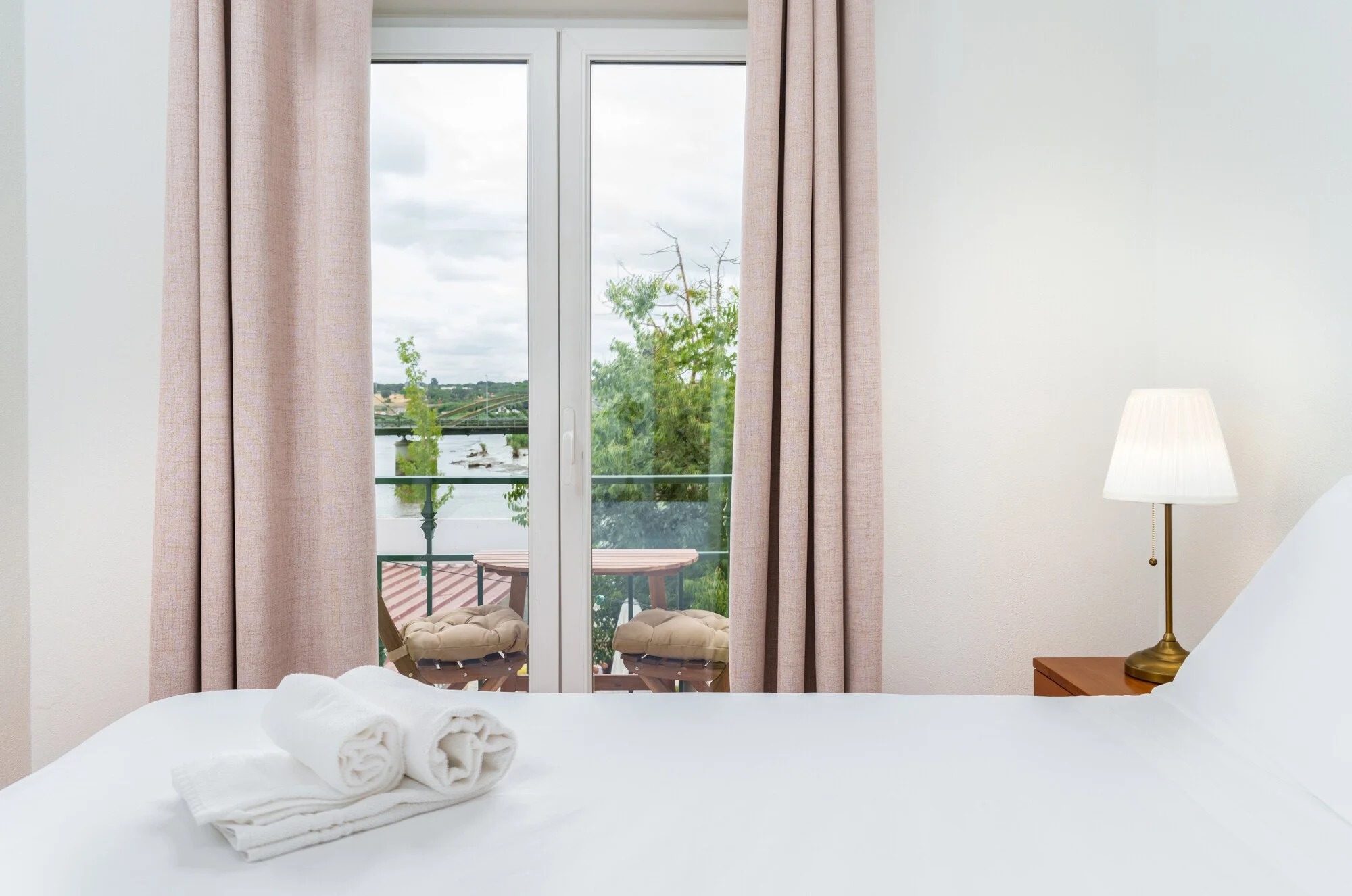 Hotel Salátia- A bedroom with a sliding glass door leading to a balcony, beige curtains, a white bed with rolled towels, a table with a white lamp, and a scenic view of trees, a river, and a bridge outside.