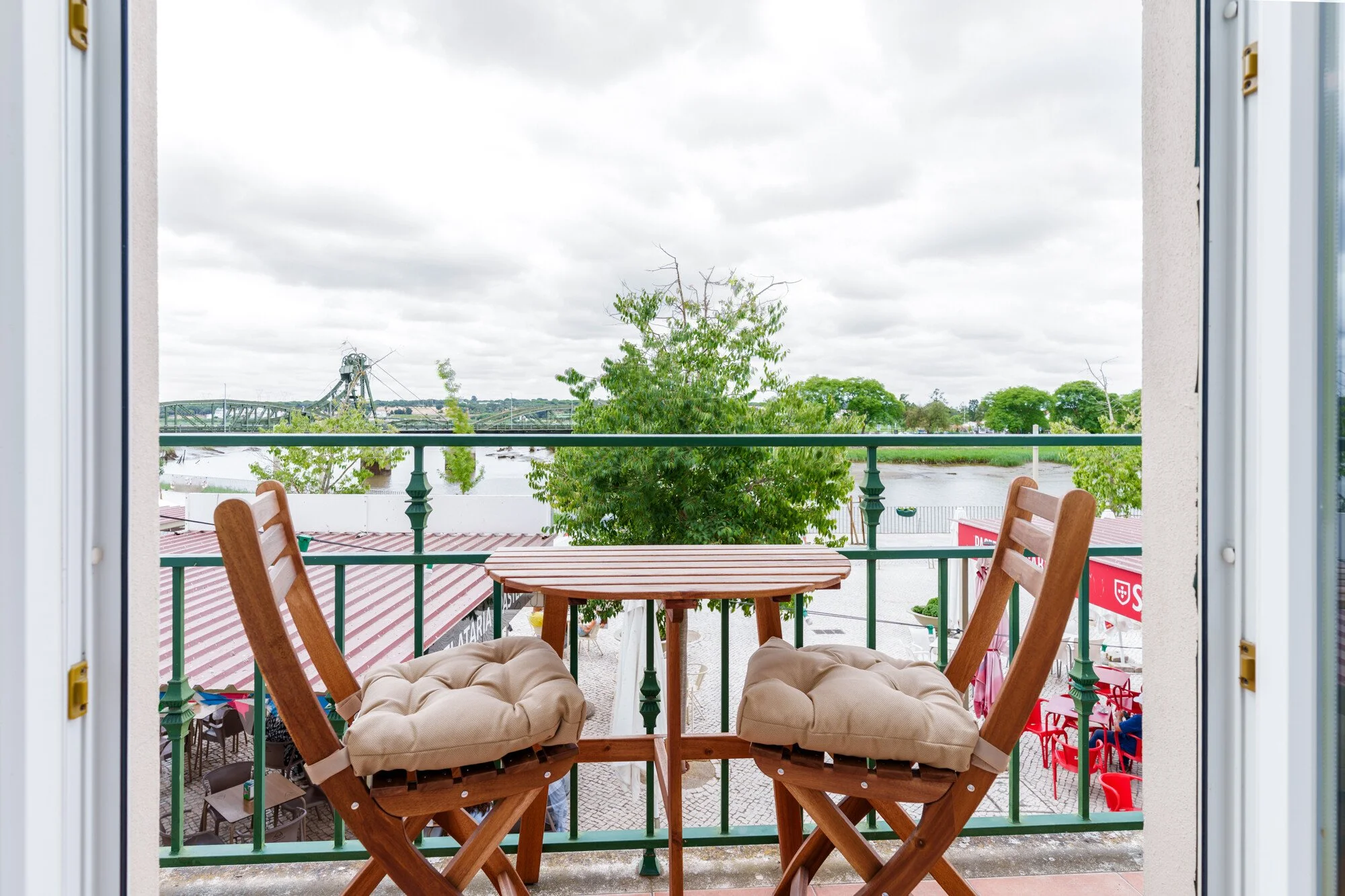 Hotel Salátia- bedroom design. Balcony view with outdoor furniture, trees, and a cloudy sky, as seen through an open window.