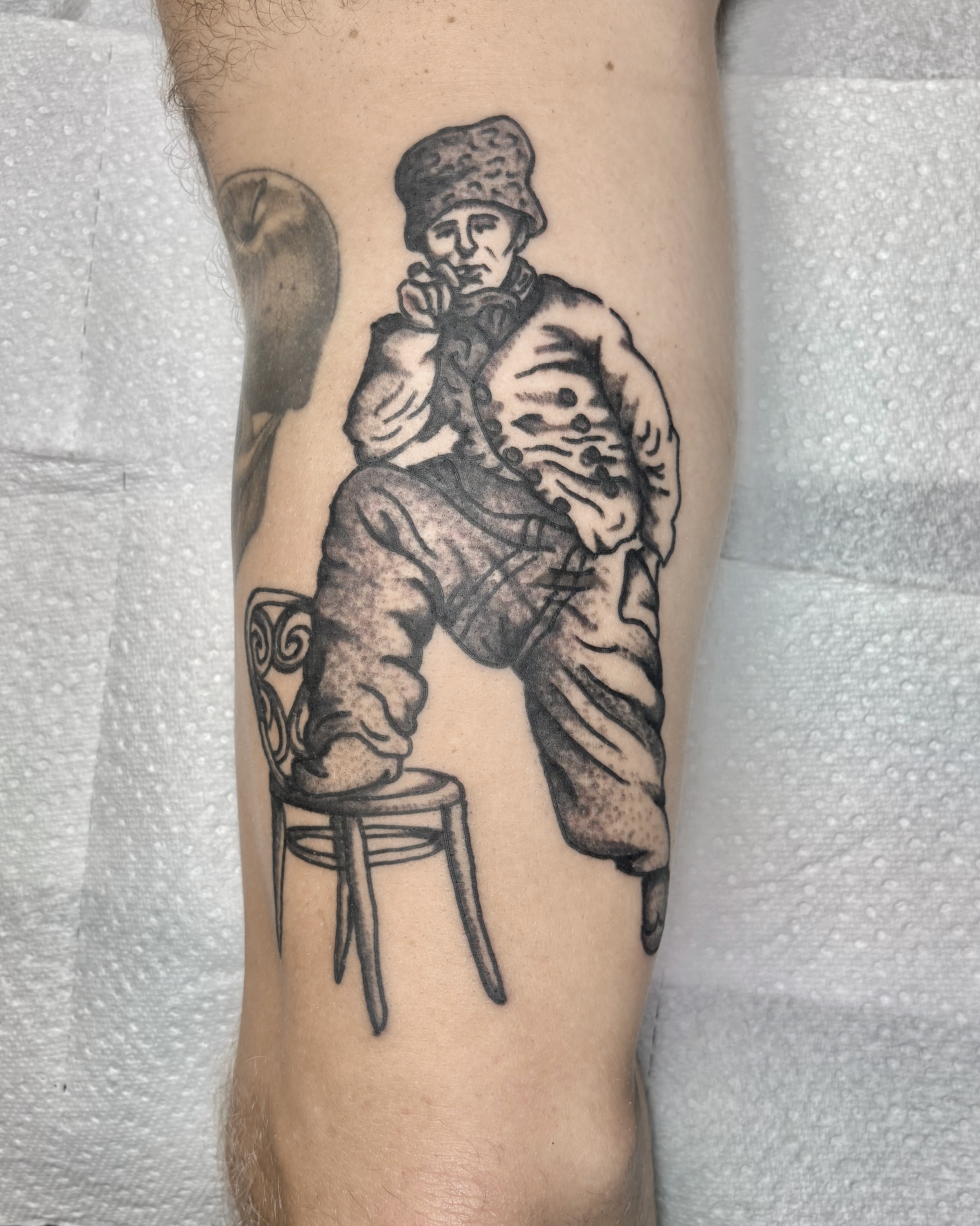 Black and gray tattoo of a man in traditional clothing, sitting on a stool with one foot on the ground and the other on the stool, holding a pipe to his mouth.