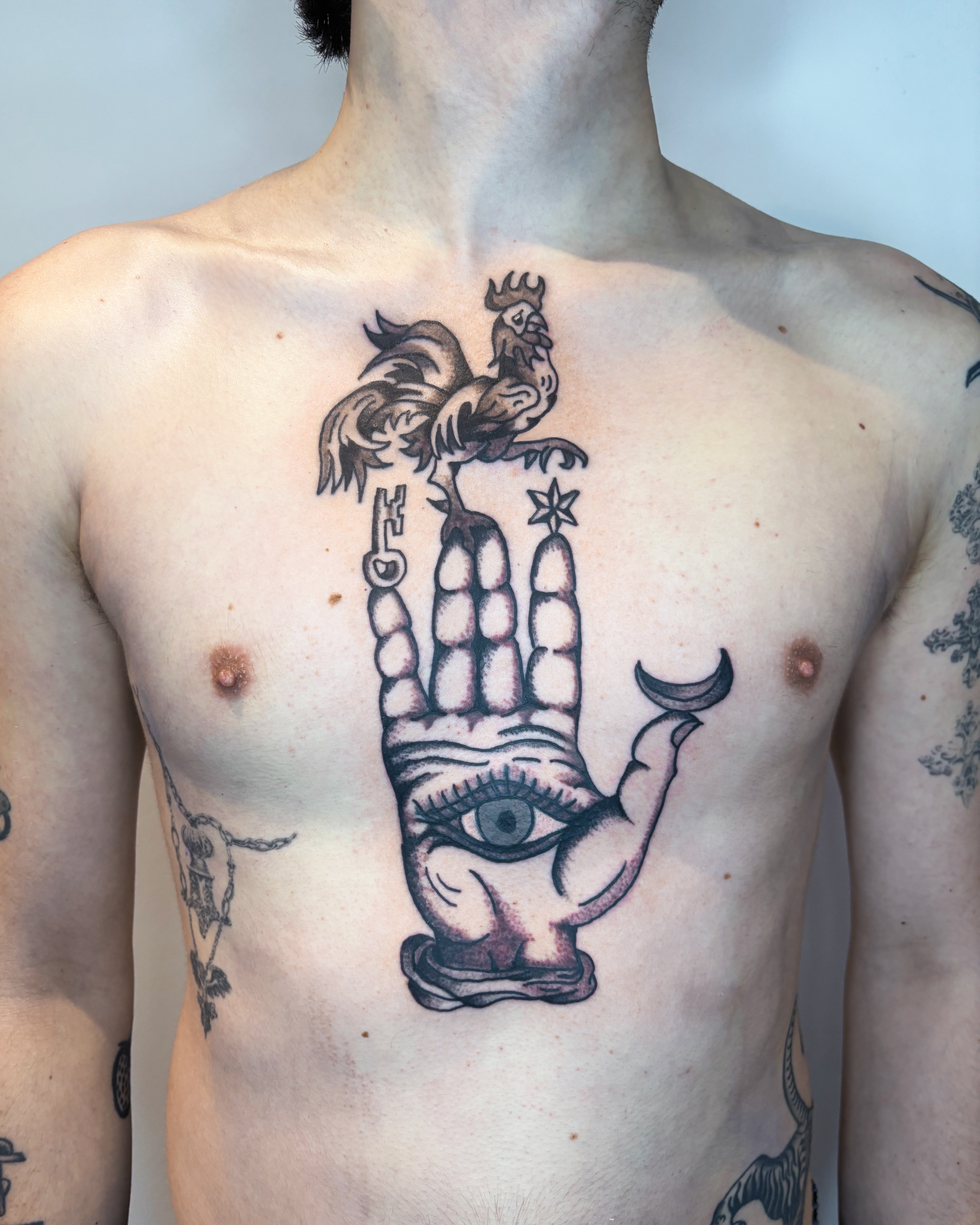 Tattoo of a large hand with an eye in the palm, a moon on the finger, a star on another finger, a rooster on the wrist, and a crescent moon on the pinky finger.
