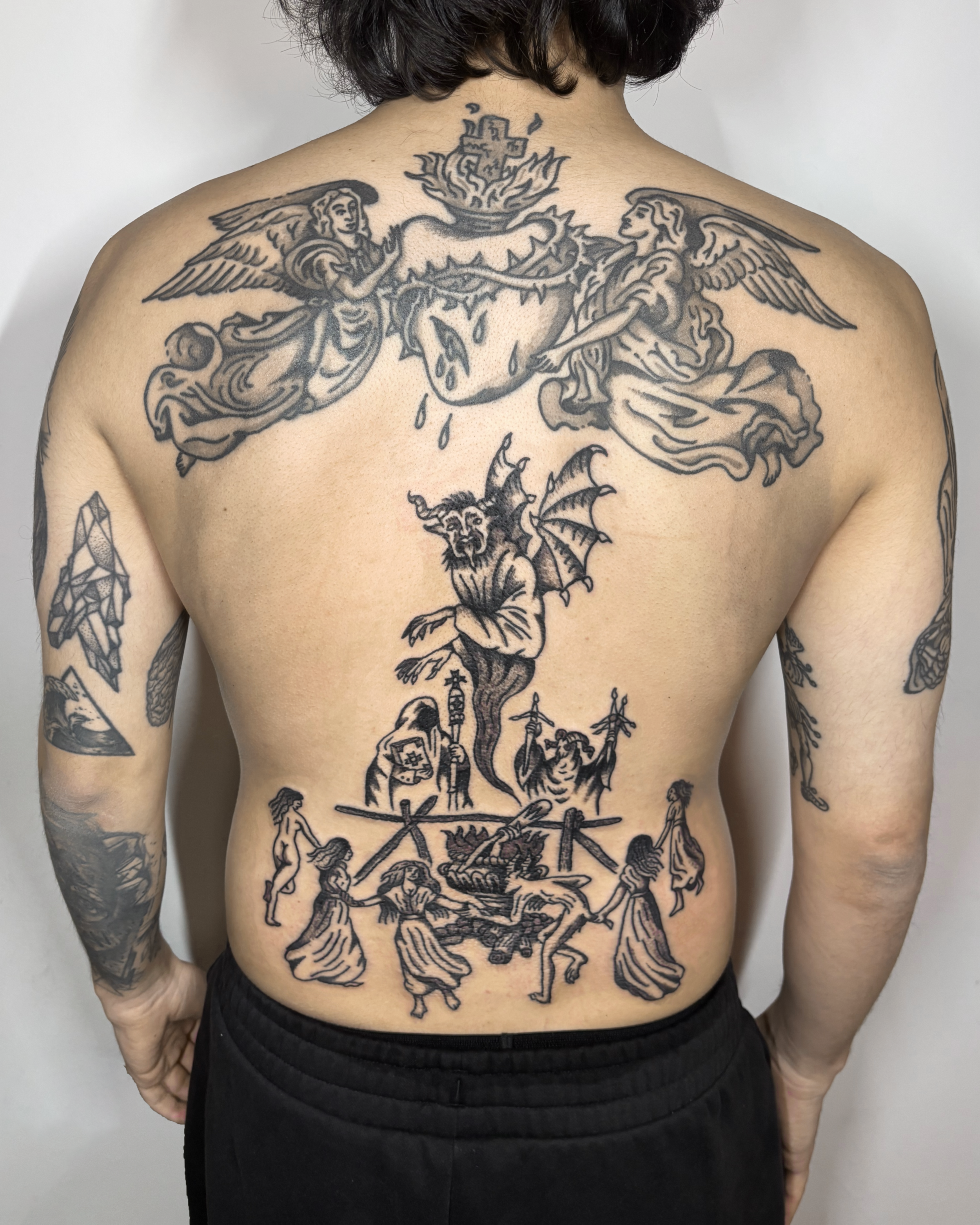 A person's back with large black line tattoos depicting mythological and fantastical scenes, including angels, a dragon, demons, and human figures.