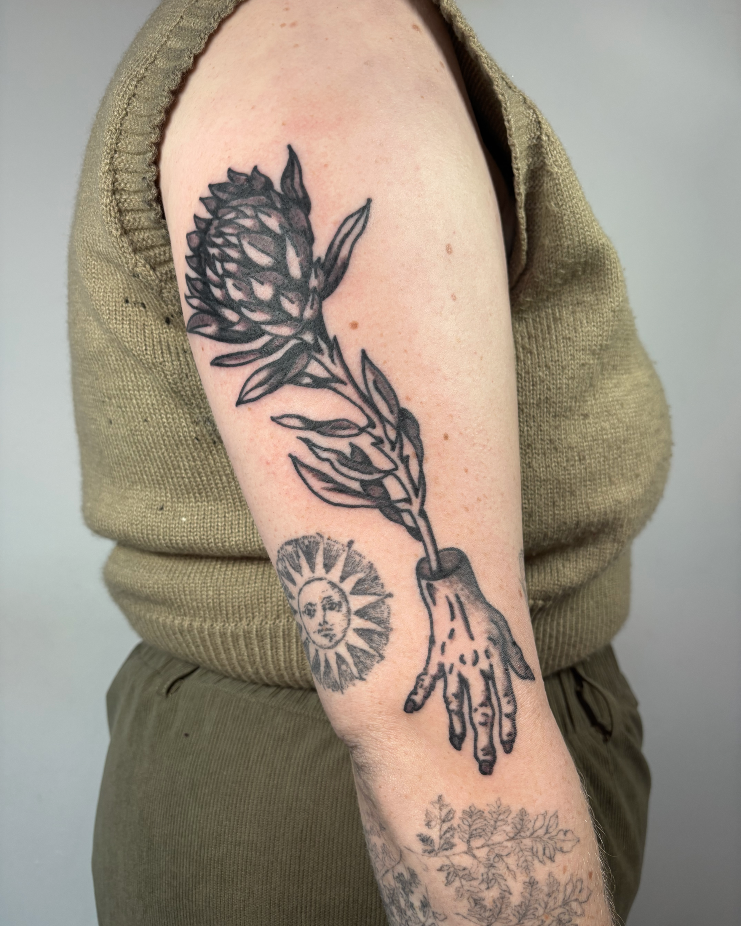 Tattoo of a hand holding a plant with a large, detailed flower, and a sun with a face on the inner arm.
