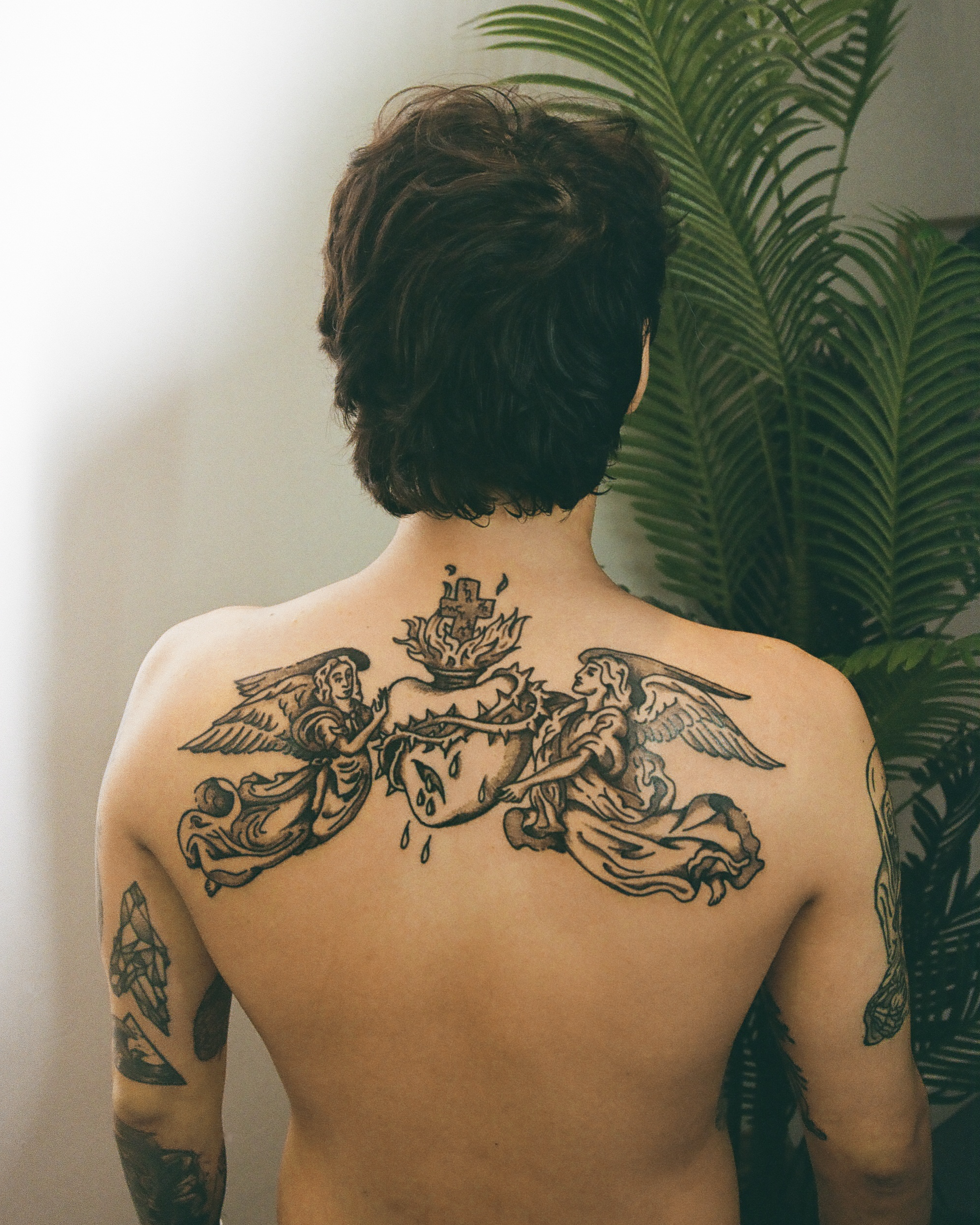 A person with dark hair and tattoos on their back and arms standing in front of green plants, facing away from the camera.