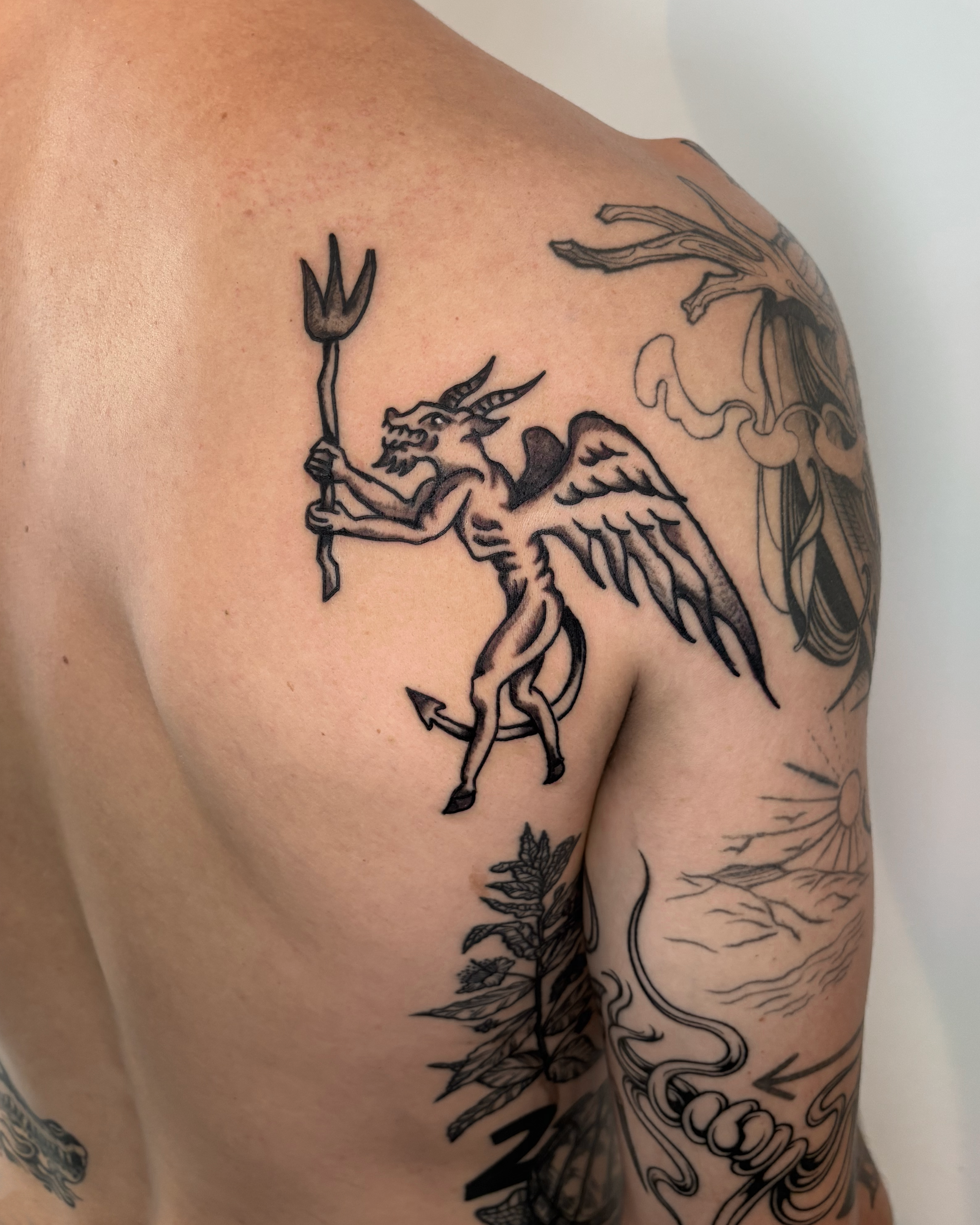 Black ink tattoo of a mythological creature with the body of a goat, wings, and a goat's head, holding a trident, on a person's upper arm.