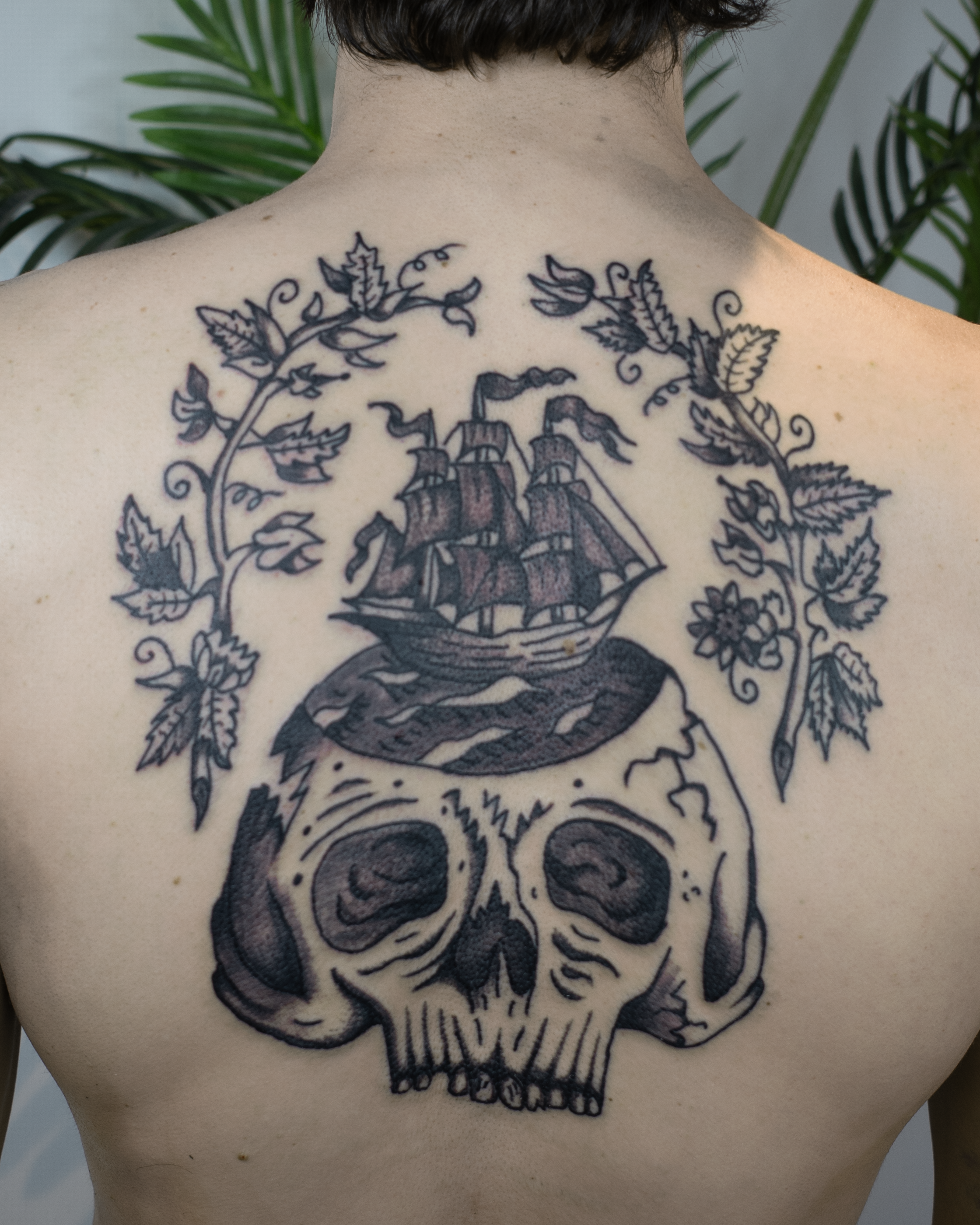 Tattoo on person's back featuring a large skull with a sailing ship on top, surrounded by leaves and floral details.