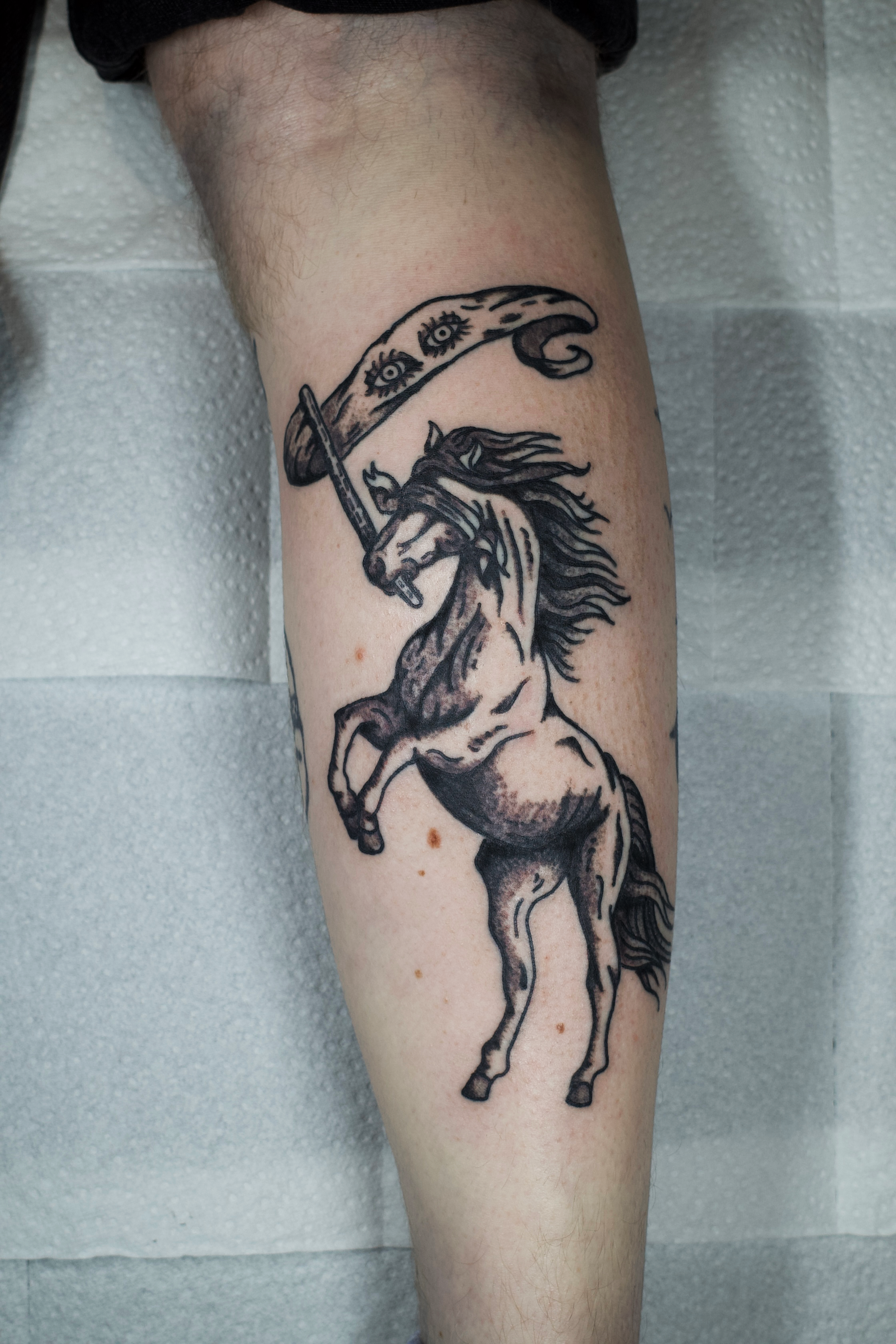Tattoo of a rearing horse with a sword in its mouth, holding a flag with eyes on it.