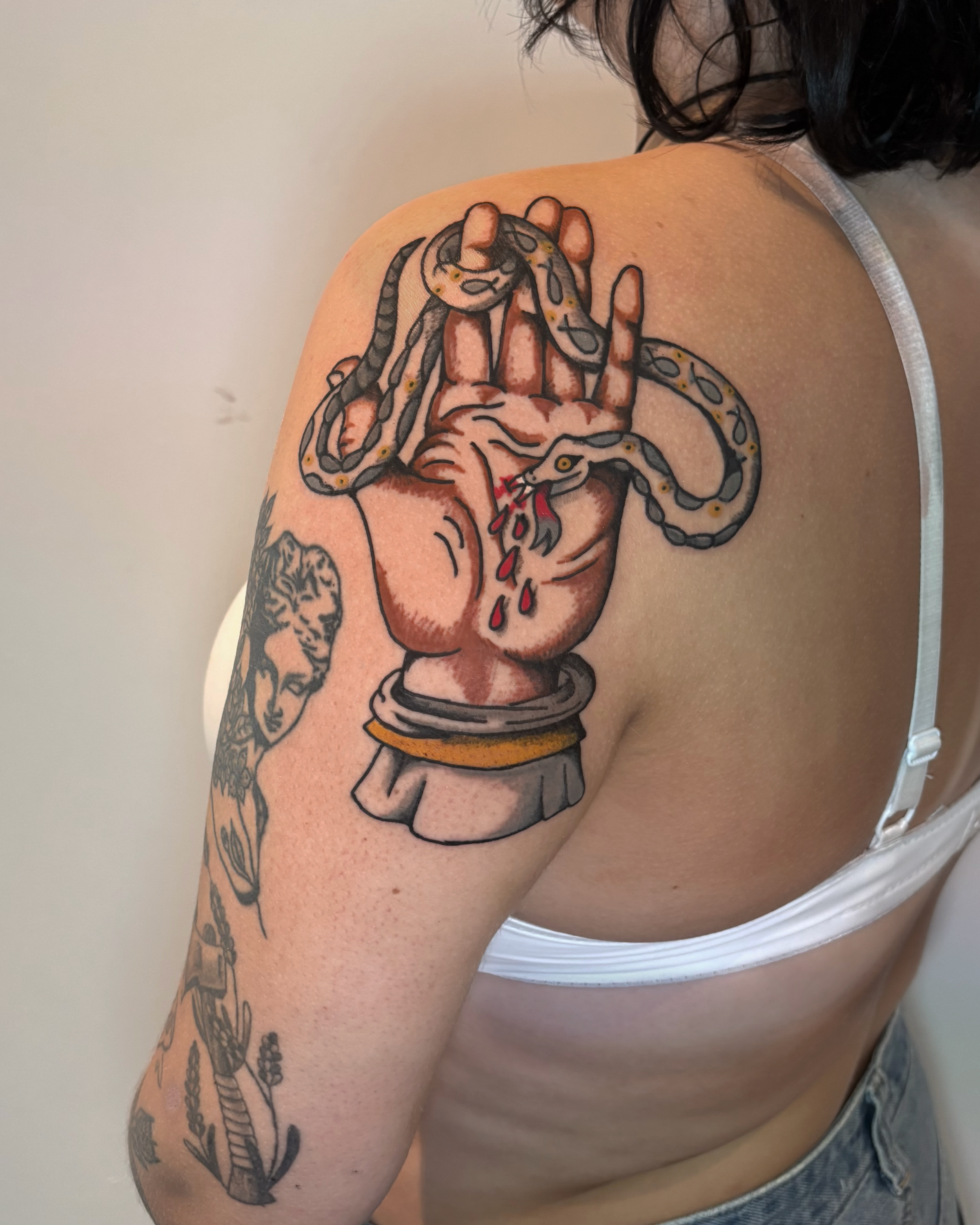 Tattoo of a hand holding a snake, with the snake's head biting and bleeding. The tattoo is on a person's shoulder and upper arm.