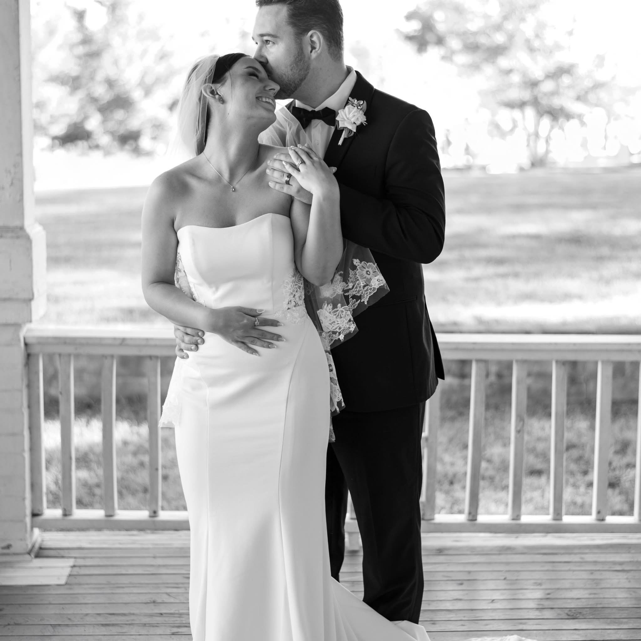 A black and white photo of a bride and groom enjoying a romantic moment. The groom is kissing the bride's forehead, and they are both smiling. The bride is wearing a strapless wedding dress, a veil, and jewelry, and the groom is in a tuxedo with a bo