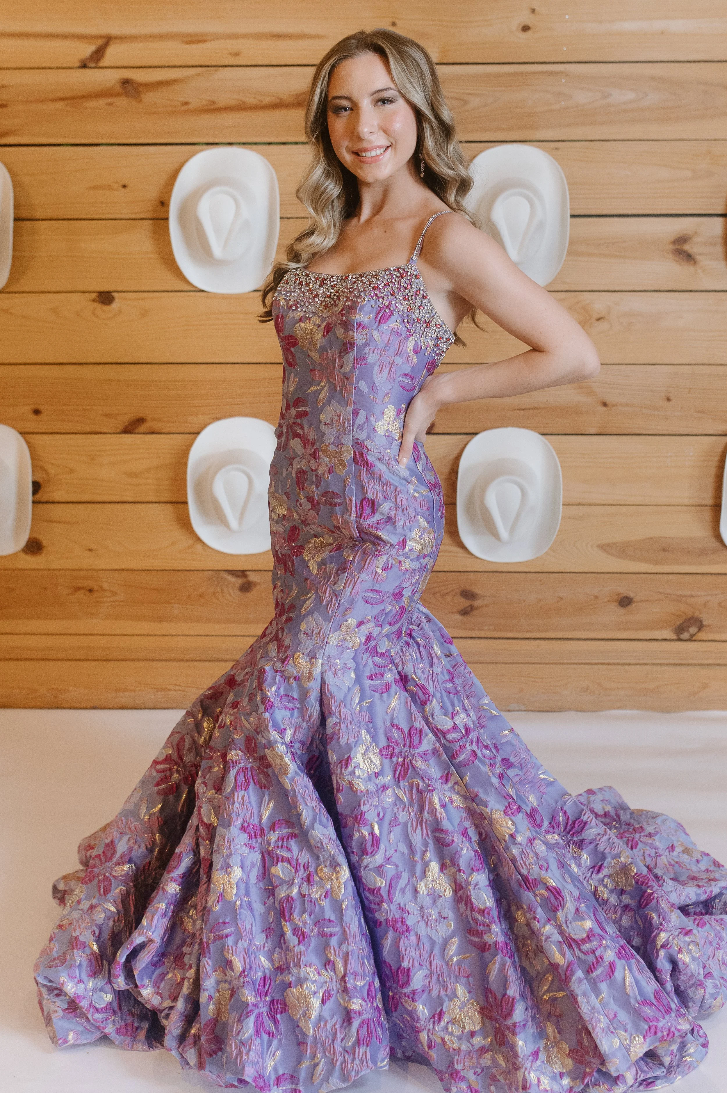 A woman in a purple floral gown with gold accents, standing in front of a wooden wall decorated with white cowboy hats. She is smiling and posing with her right hand on her hip.