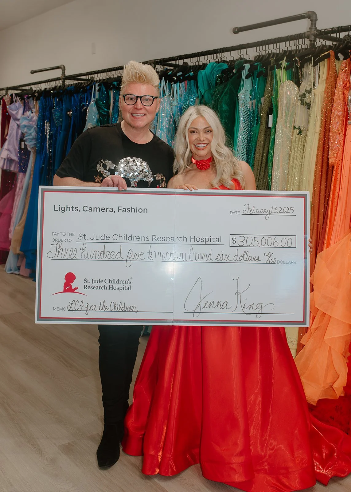 Two people, a man and a woman, stand in front of a colorful display of dresses, holding a large check made out to St. Jude Children's Research Hospital for over 305,000 dollars. The man has light-colored hair and glasses, wearing a black T-shirt, and