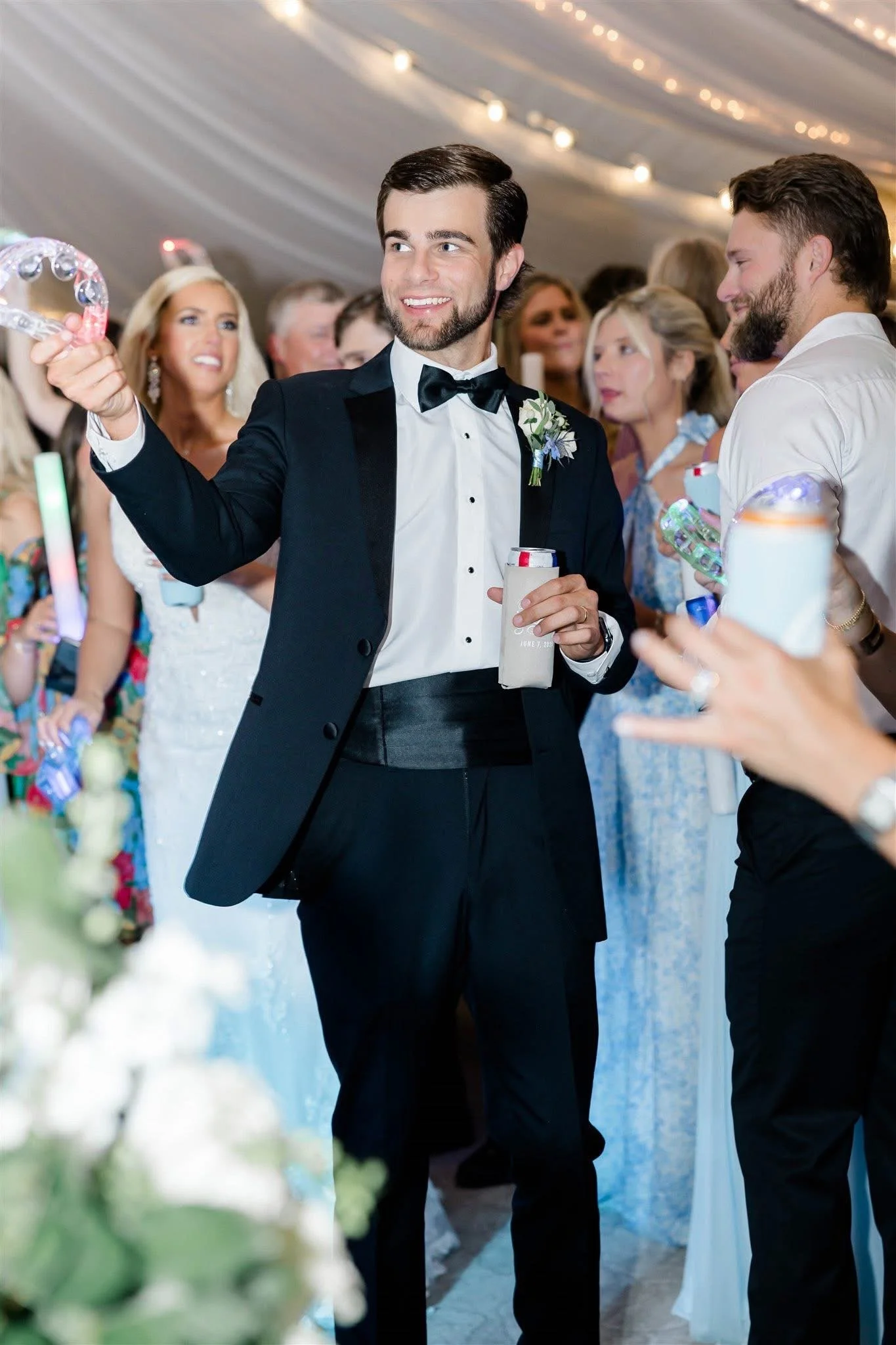 A man in a tuxedo holding a tambourine at a wedding reception surrounded by guests.