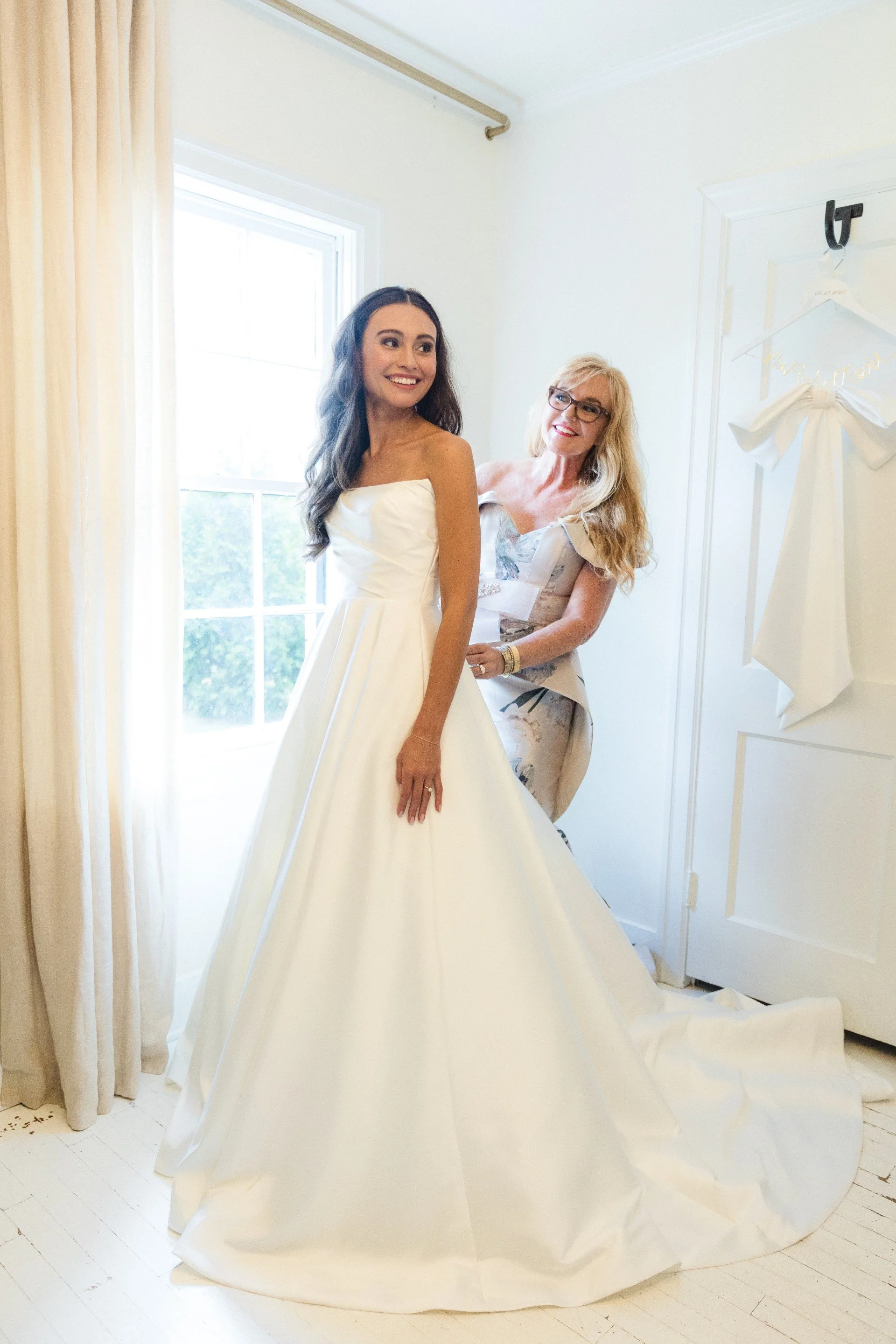 A bride in a white wedding dress standing by a window with sunlight, smiling, while an older woman helps her with the dress, in a bright room with white walls and curtains.