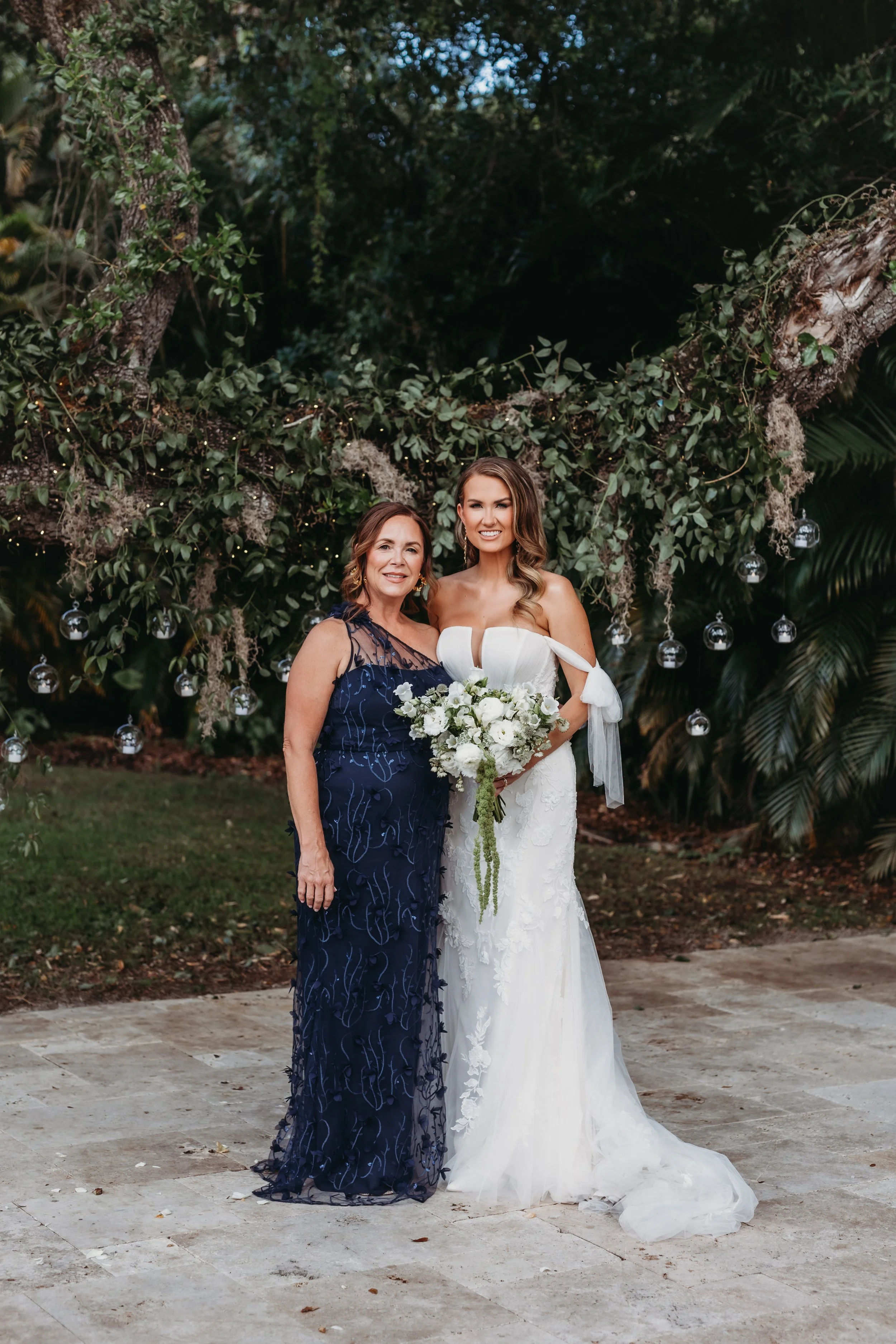 A bride in a white wedding dress holding a bouquet of white flowers standing next to a woman in a navy blue dress, outdoors beneath a large tree decorated with hanging glass ornaments and string lights.