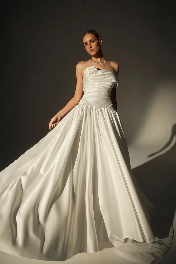 A woman wearing a white strapless wedding gown with a fitted bodice and a full, flowing skirt, standing against a dark background.