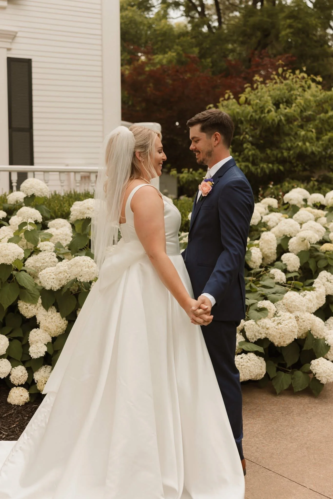 A newlywed couple holding hands and smiling at each other outdoors, surrounded by white hydrangea flowers and trees with colorful fall foliage.