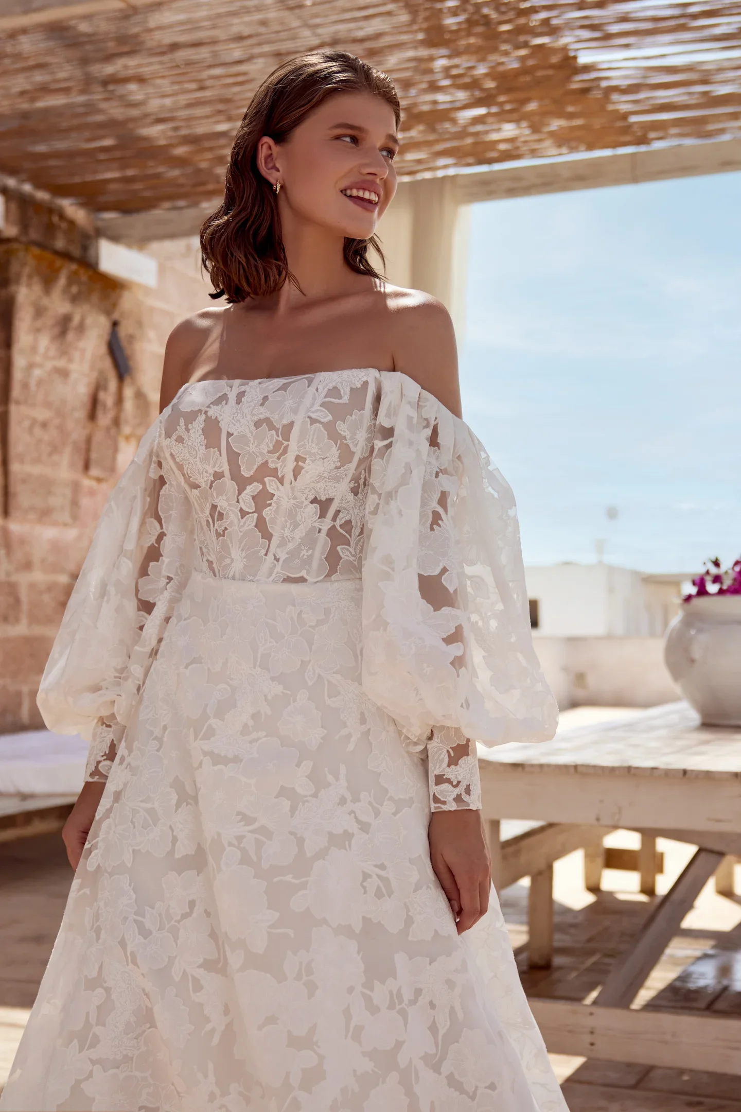 A young woman in a white lace wedding dress with off-shoulder sleeves standing outdoors under a wooden canopy, with the ocean and sky in the background.