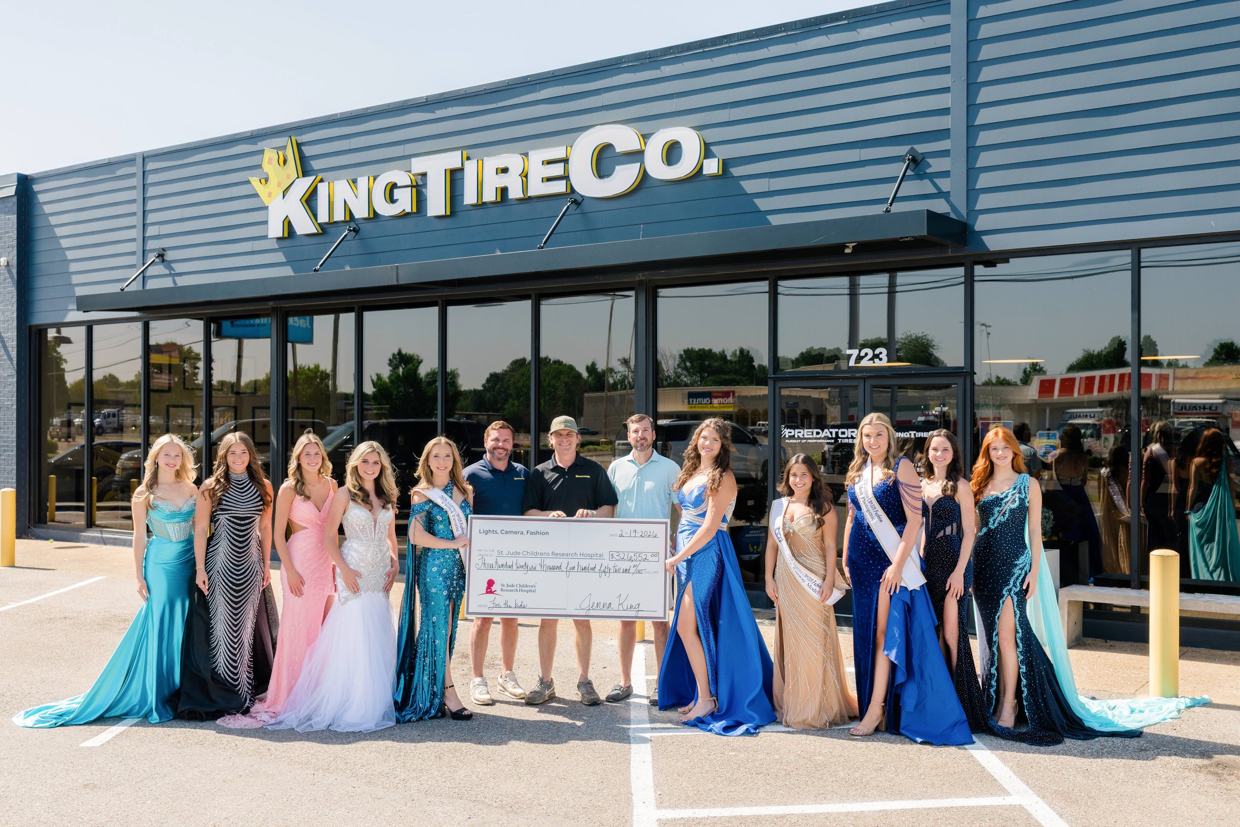 A group of people, including women in colorful evening gowns and men, standing outside a store called King Tire Co., holding a large check for over three hundred thousand dollars for a children's research hospital, with a building in the background.