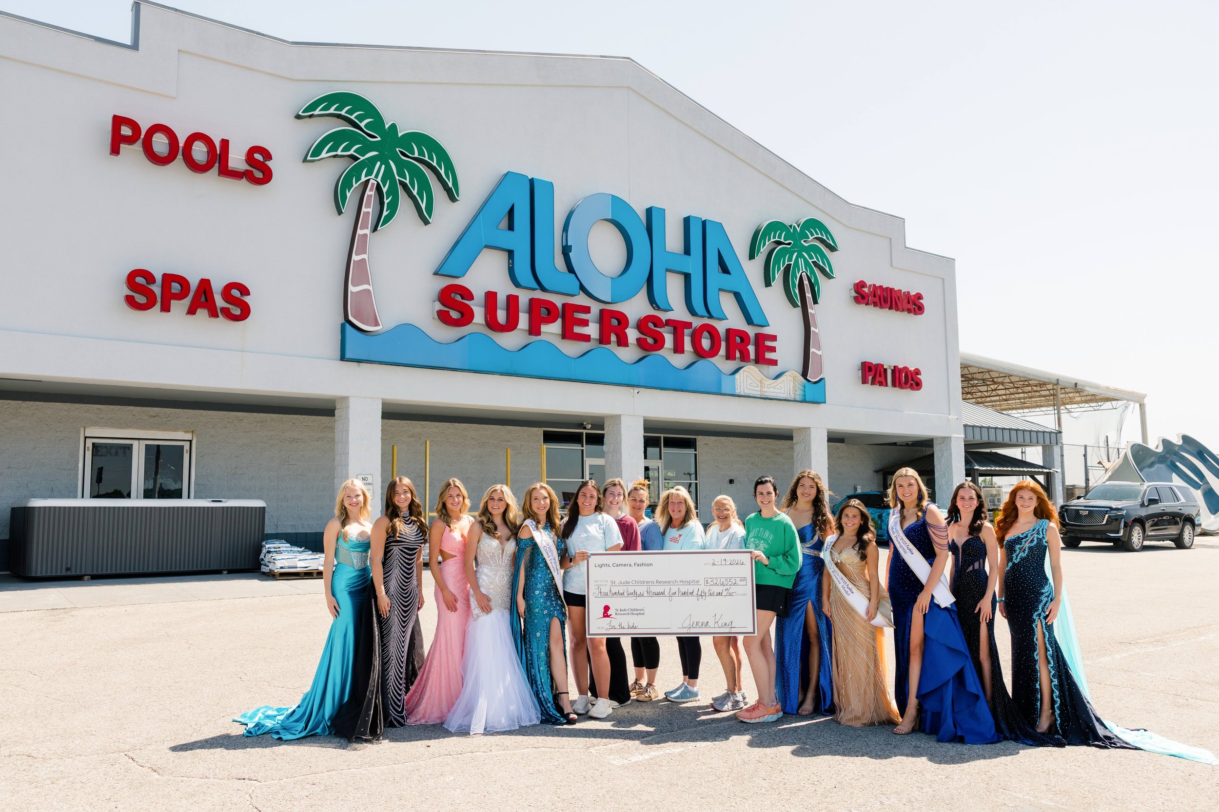A group of women in colorful dresses standing in front of an Aloha Superstore sign with palm trees and additional store signs for pools, spas, saunas, and patios. They are holding a large check and smiling at the camera.