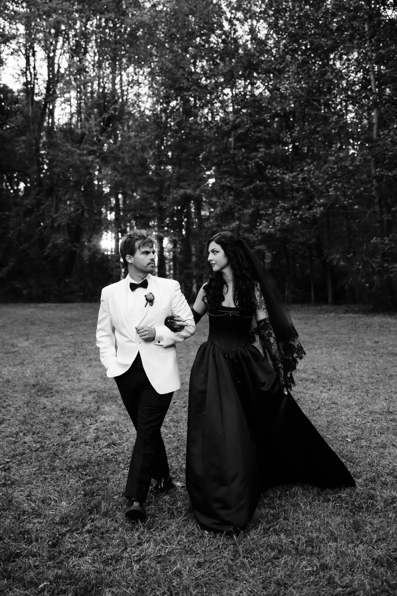A black and white photo of a man in a white tuxedo and a woman in a black gown walking outdoors on grass, surrounded by trees, as the sun sets behind the trees.