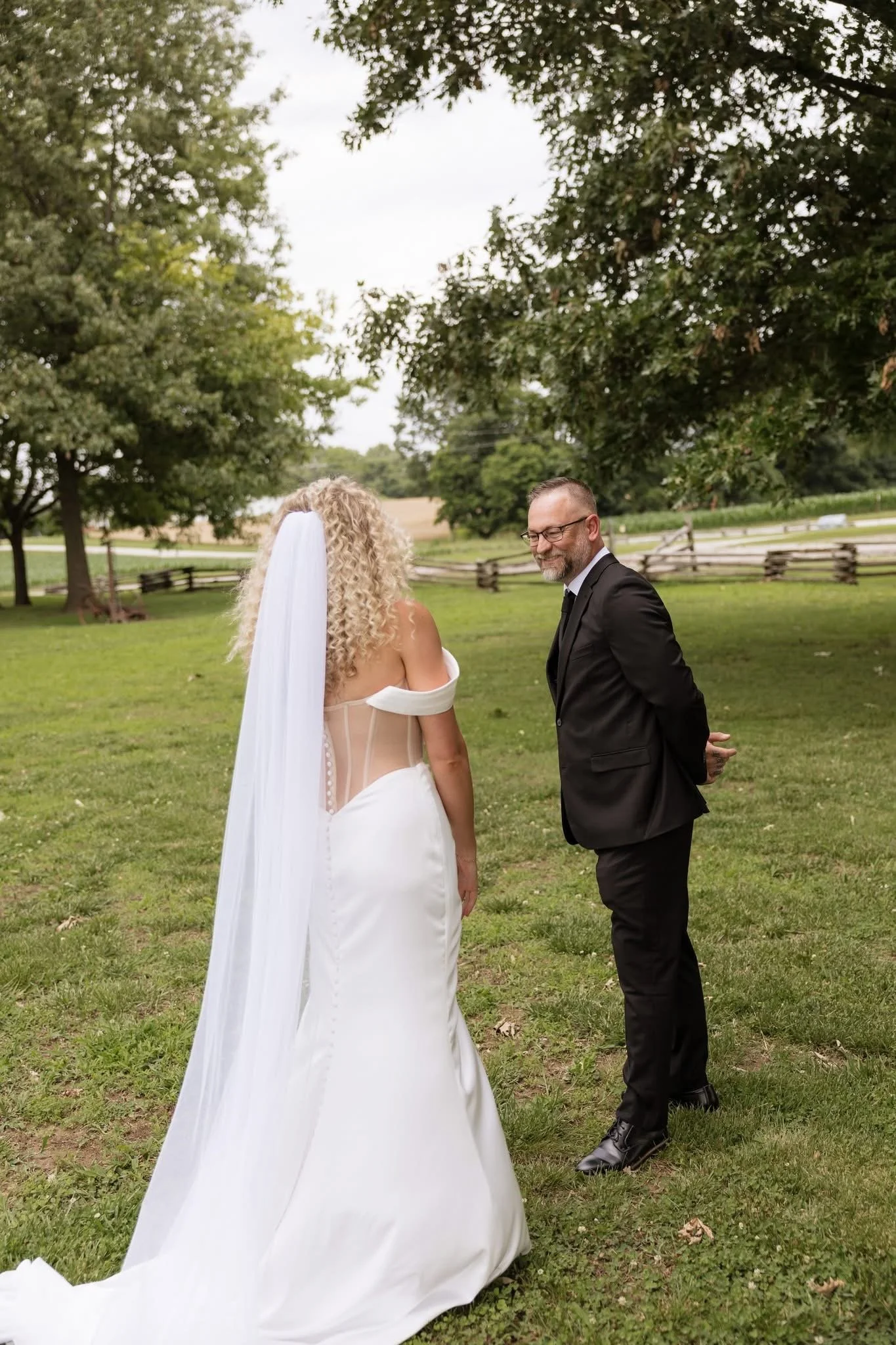 A bride with curly blonde hair in a white wedding dress and veil standing outdoors, facing a groom in a black suit, in a grassy park with trees and a wooden fence in the background, during daytime.