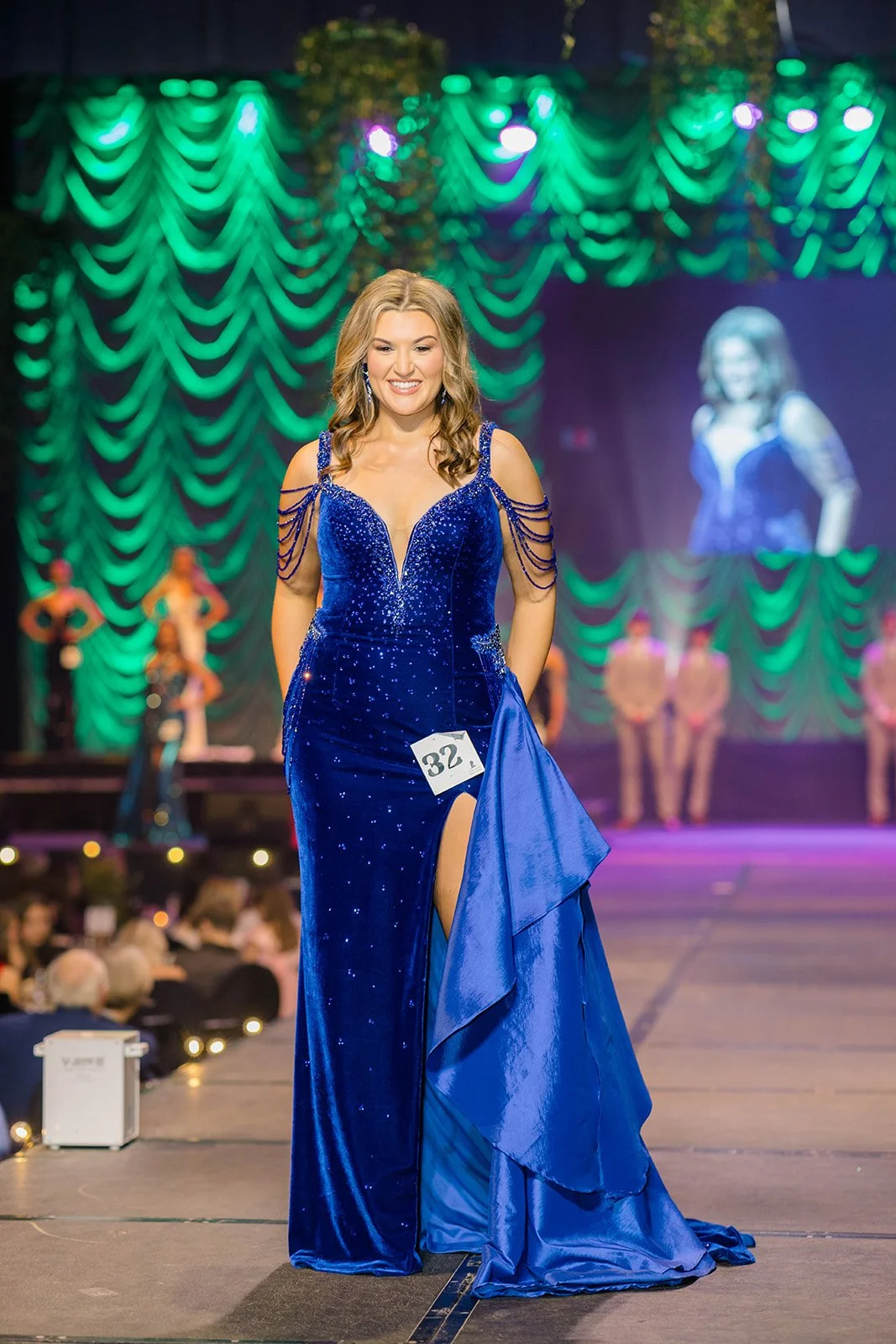 Miss USA contestant wearing a royal blue gown with sparkles and cut-out shoulders, walking on stage with a smile during a pageant event. She has a number 38 badge attached to her dress. The background features green curtains with purple and green stage lighting, with other contestants visible behind her.