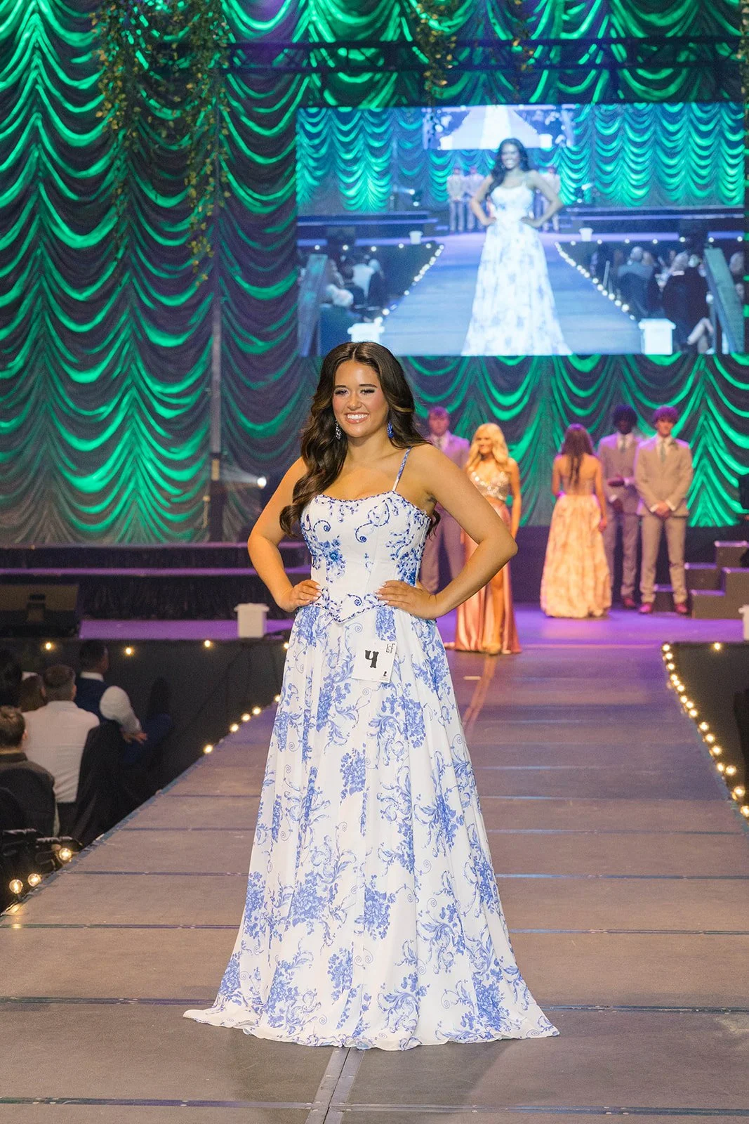 A young woman in a white and blue floral dress on stage during a beauty pageant or fashion show, with a large screen behind her displaying her walking on the runway. Other contestants are visible in the background, and the stage is decorated with green curtains and string lights.