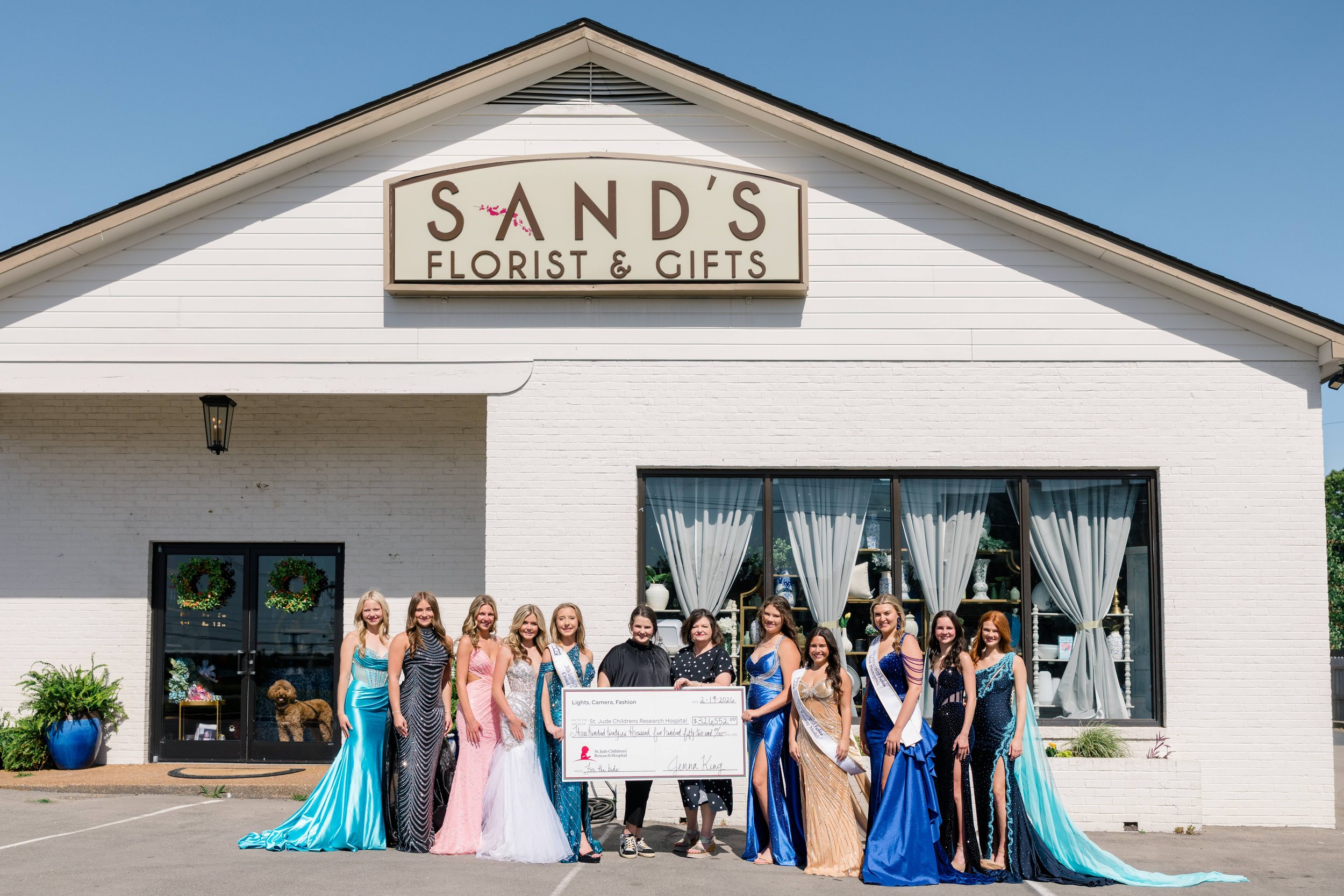 Group of women in evening gowns standing in front of a white building with a sign that reads 'SAND'S Florist & Gifts.' They are smiling and holding a large check.