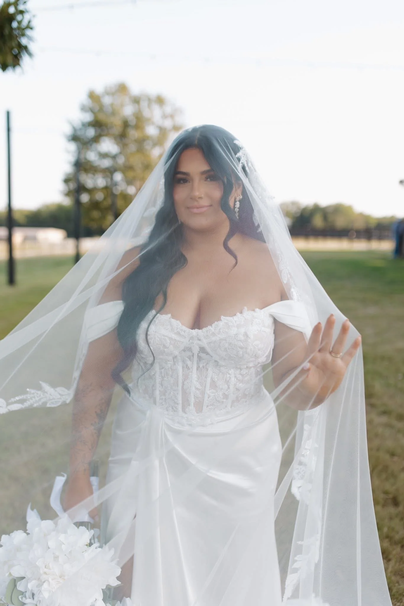 A bride in a white wedding dress with lace detail, a veil, and long dark hair standing outdoors in a park-like setting with trees and grass.