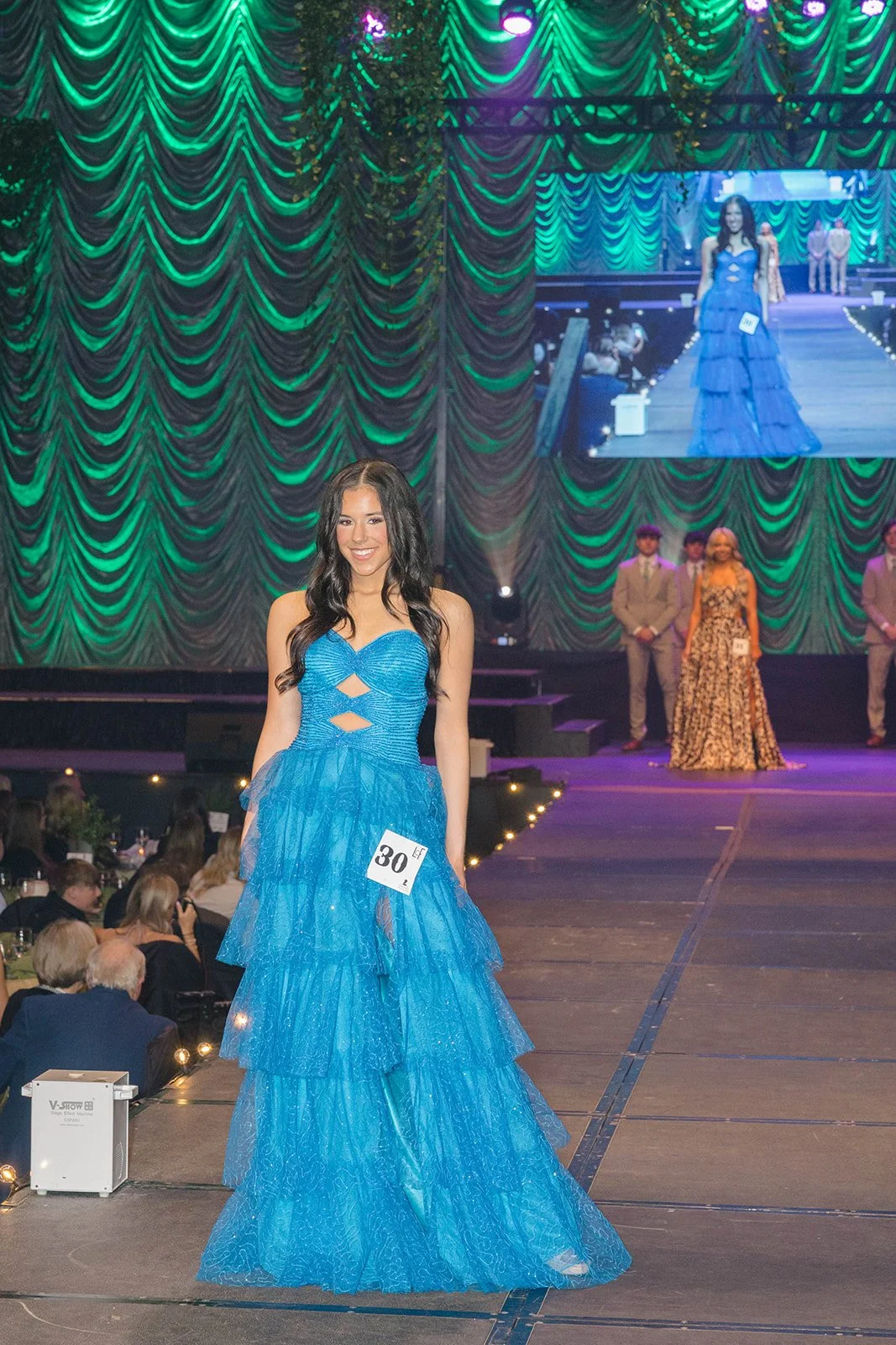 A woman wearing a bright blue evening gown and a contestant number 30 on it, standing on a stage at a fashion or beauty competition. She is smiling and has long dark hair. The stage features a large draped green curtain and a big screen projecting her image in the background. Audience members are seated at tables in the foreground.