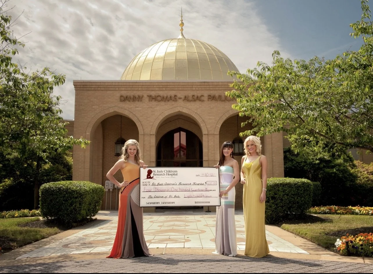 Three women in elegant dresses standing in front of a building with a gold dome, holding a large check from St. Jude Children's Research Hospital.