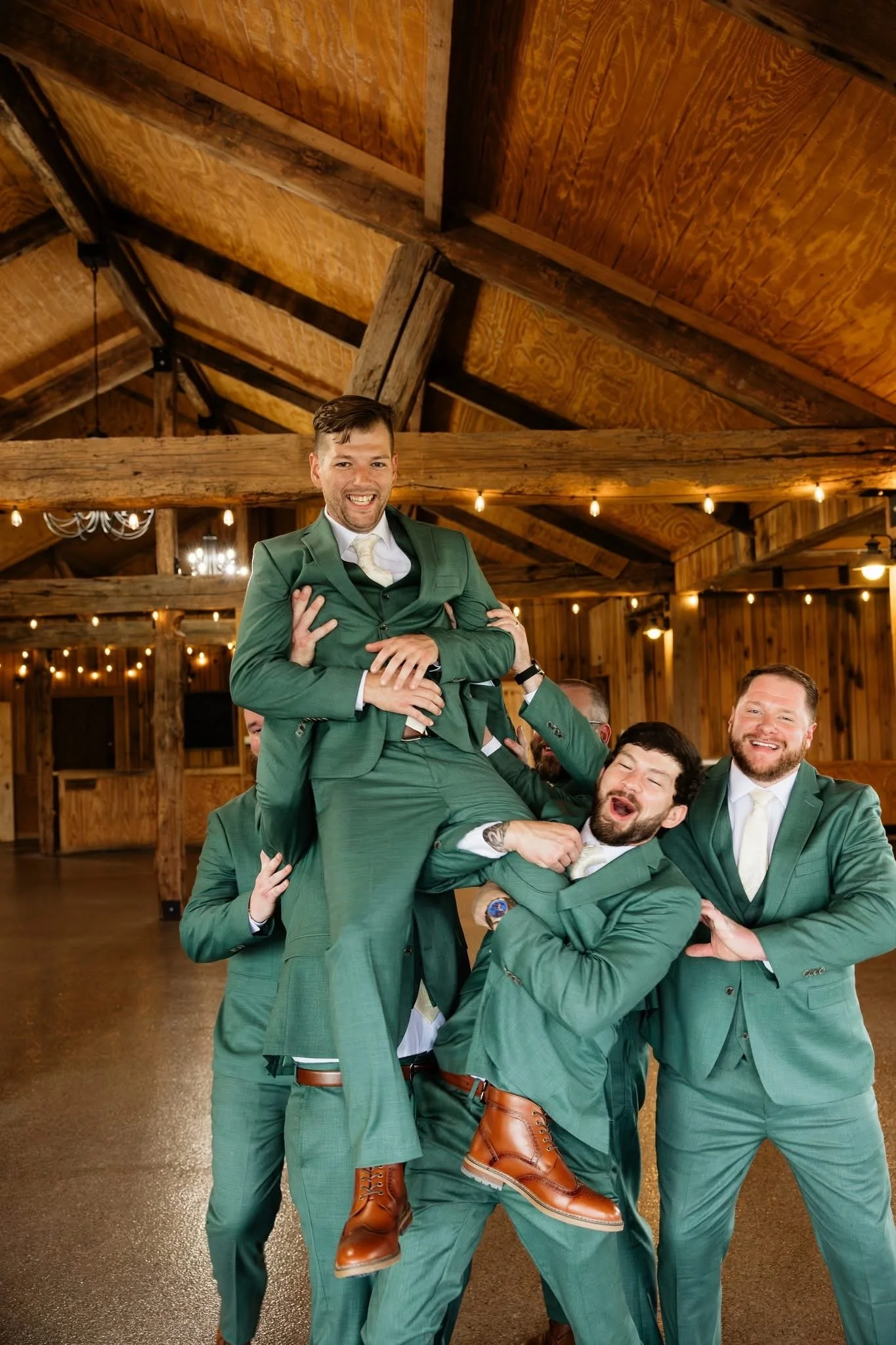 A group of men dressed in matching green suits celebrating at an indoor event, with one man being lifted by others. The background features wooden beams and warm lighting.