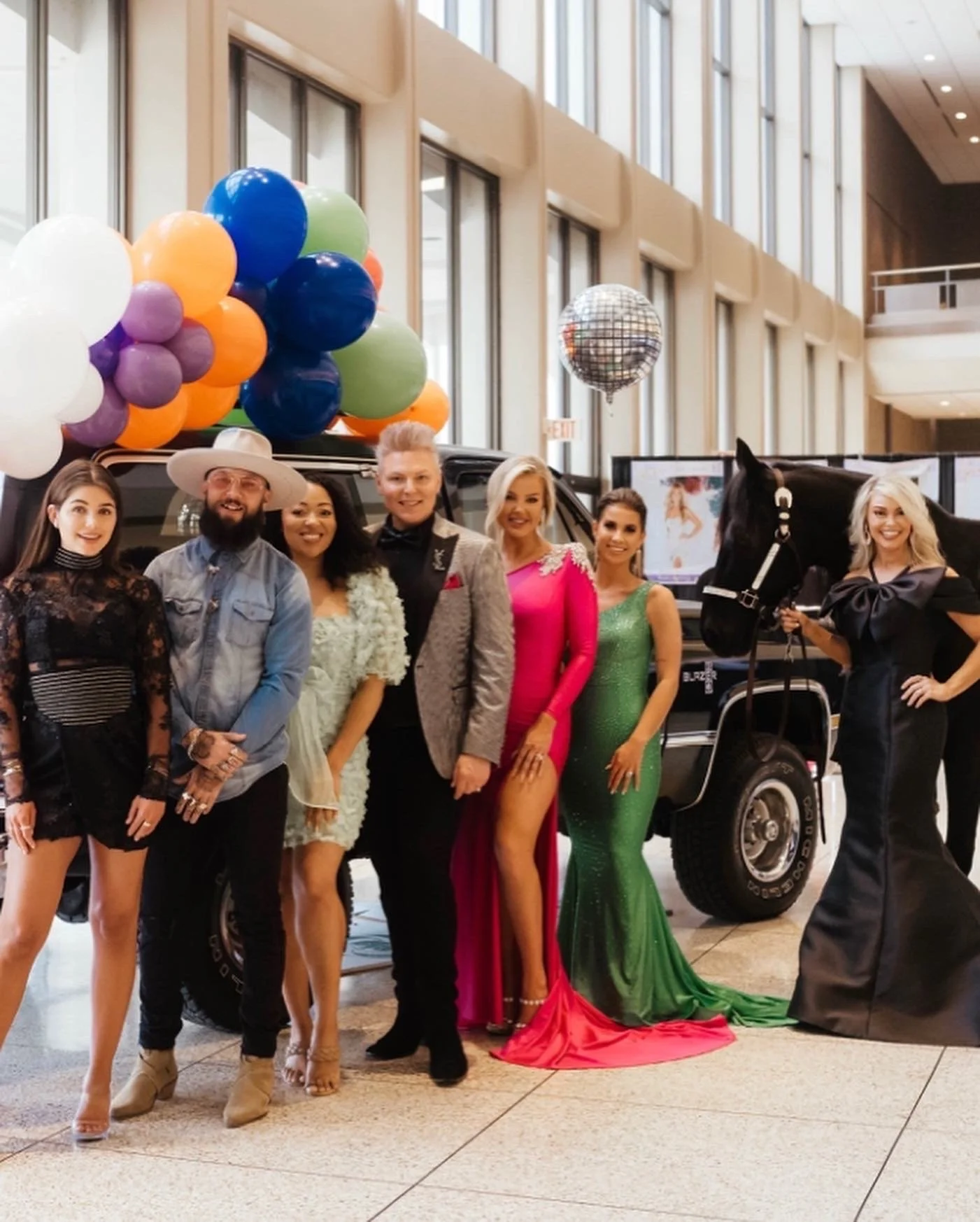 A group of seven people dressed in colorful and fashionable outfits posing in front of a black vehicle with balloons and a disco ball decoration in an indoor event space.