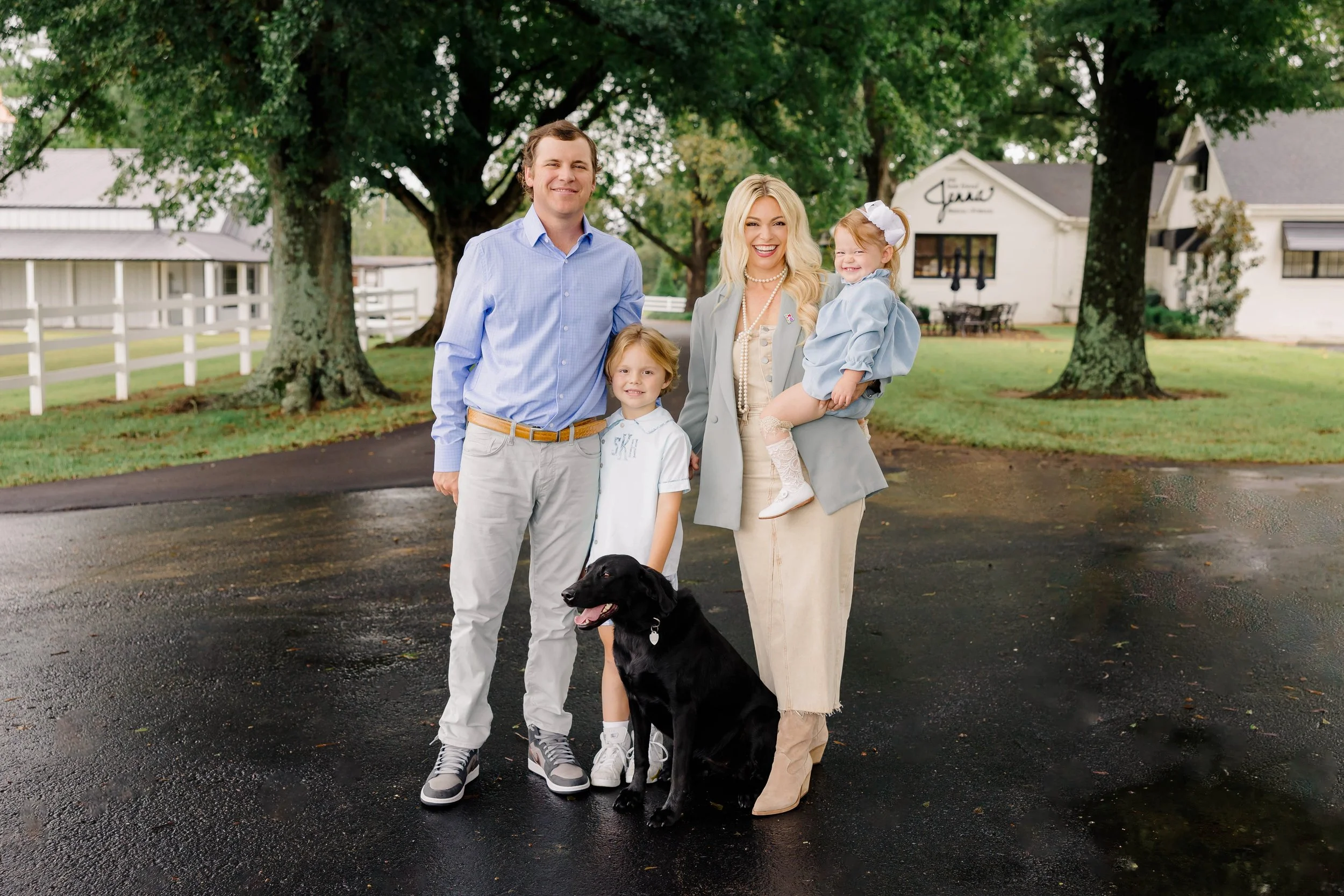 A family of four with a black lab dog standing on a wet driveway in front of a farm or ranch with trees and white buildings, smiling at the camera.