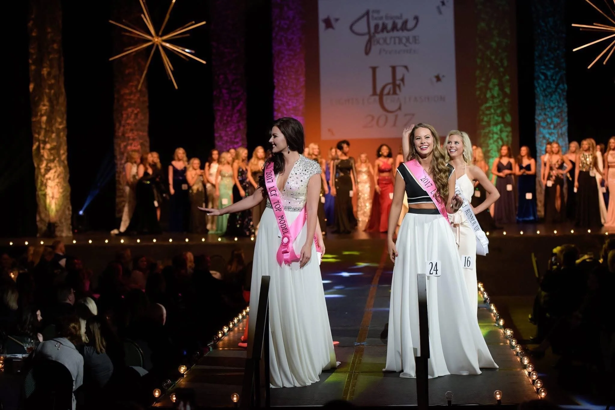 Beauty pageant contestants on stage wearing sashes and evening gowns, with a colorful backdrop and audience in the foreground.