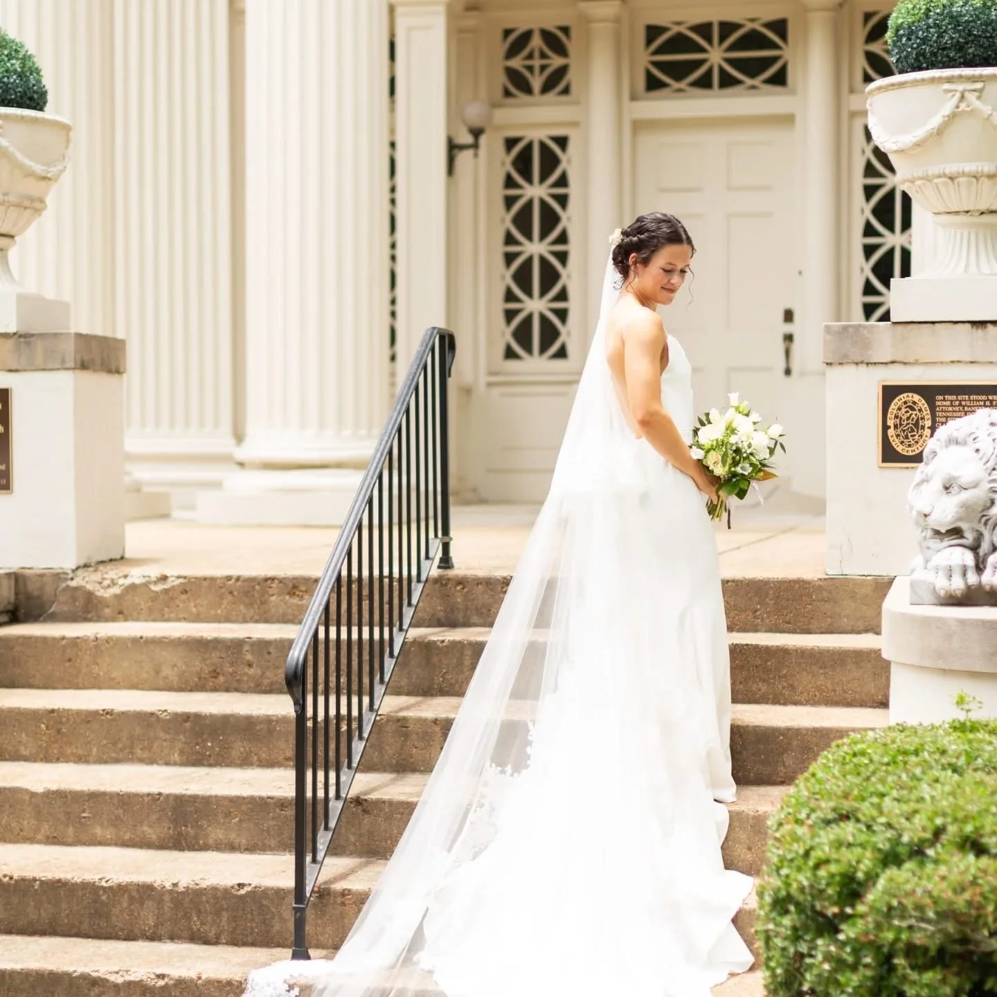 Bridal gown bride holding a bouquet of white flowers on the steps outside a building with white columns and black railings.
