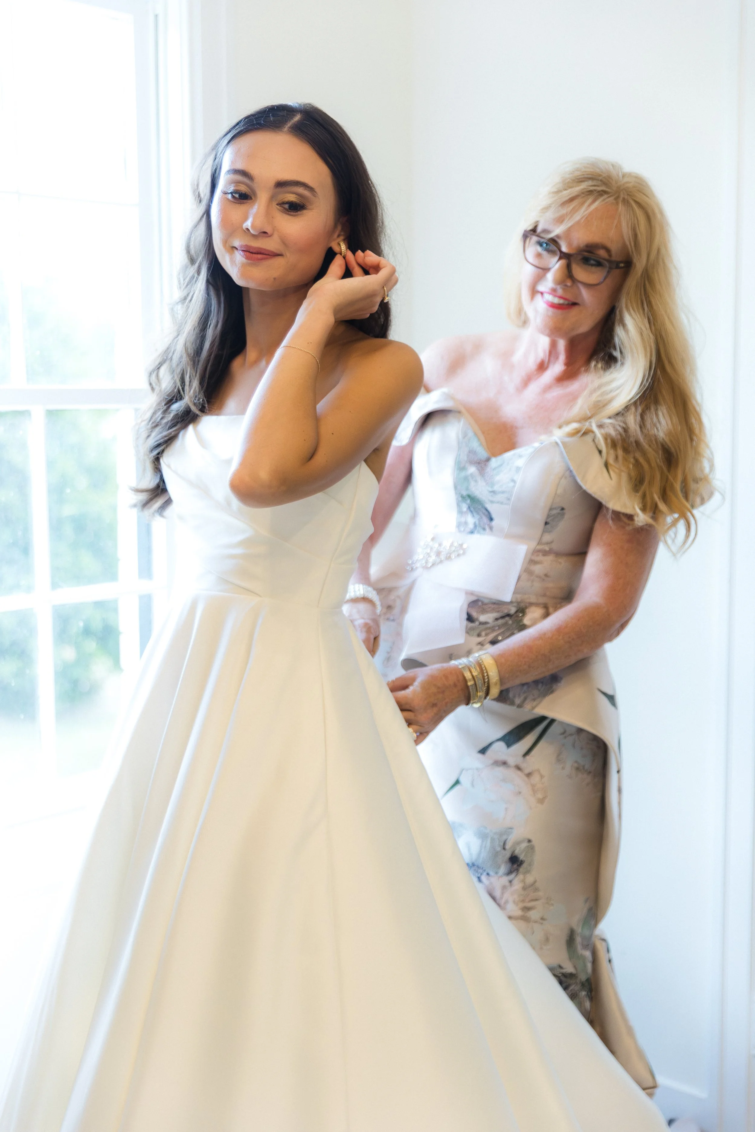 A woman in a wedding dress putting on an earring while another woman helps her, in a bright room with a large window.