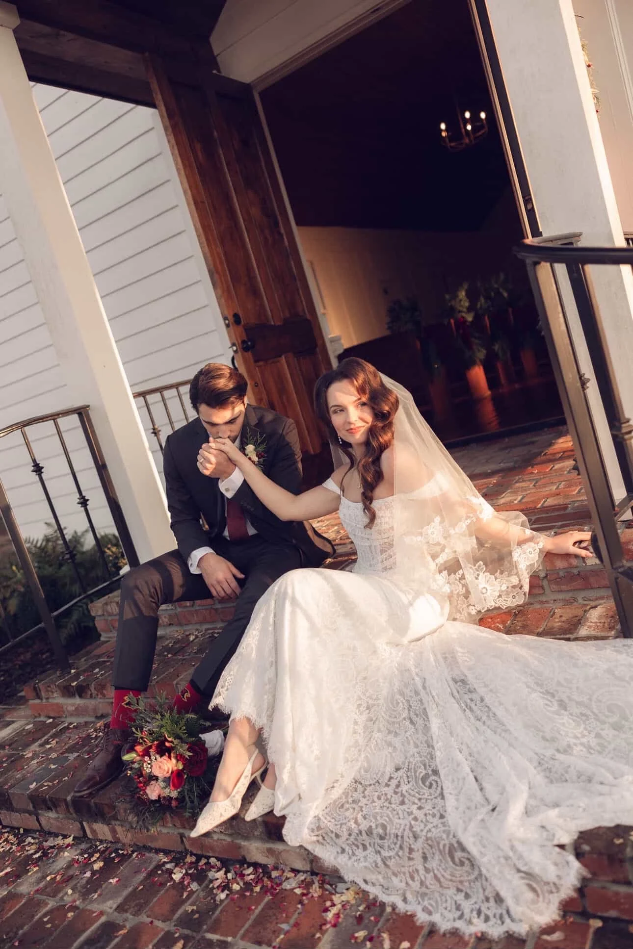 A bride in a lace wedding gown and a groom in a suit sitting on brick steps outside a house, with flowers and confetti around them.