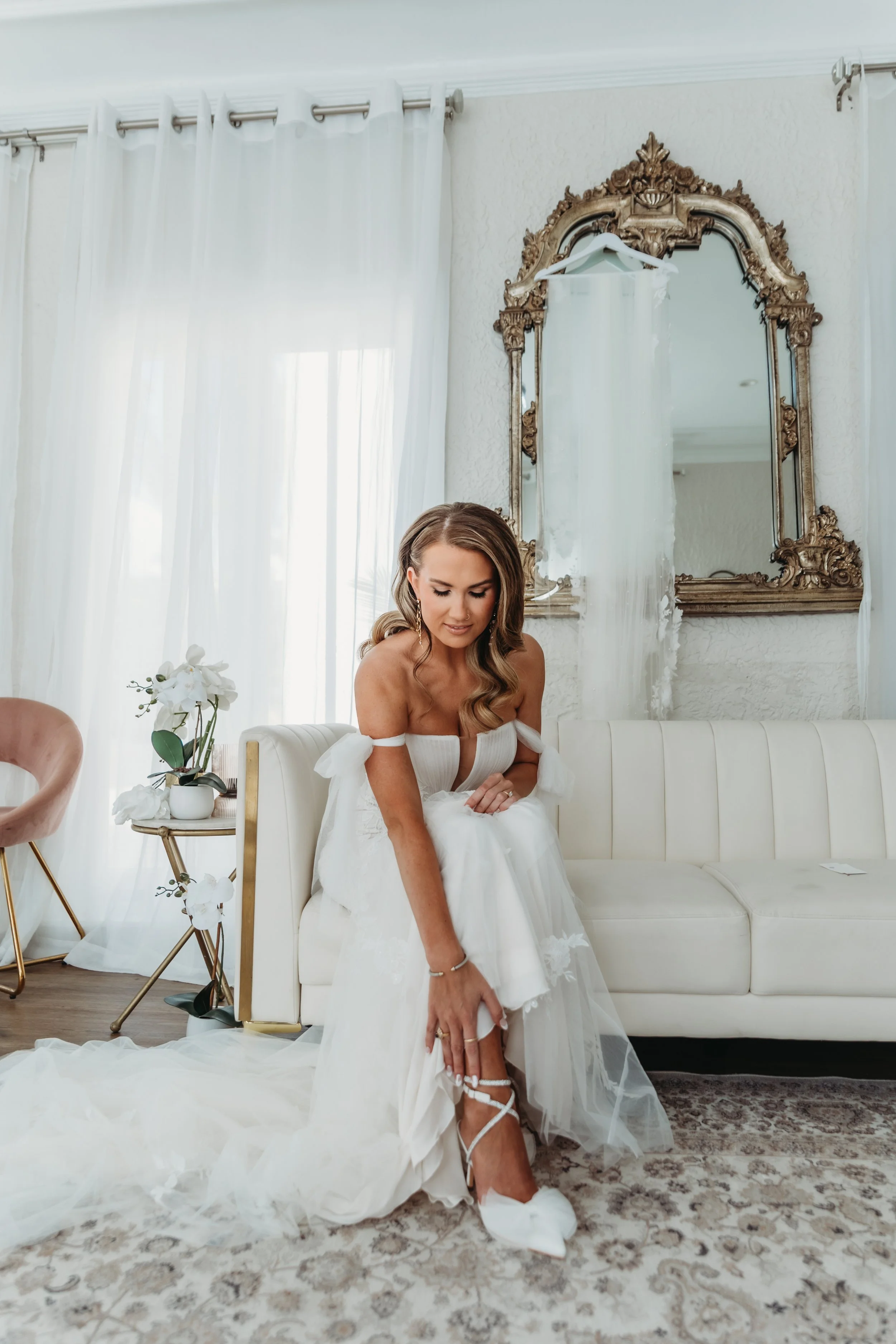 A bride in a white wedding dress is sitting on a white couch, adjusting her shoe, with a large ornate mirror and white curtains behind her in a bright, elegant room.