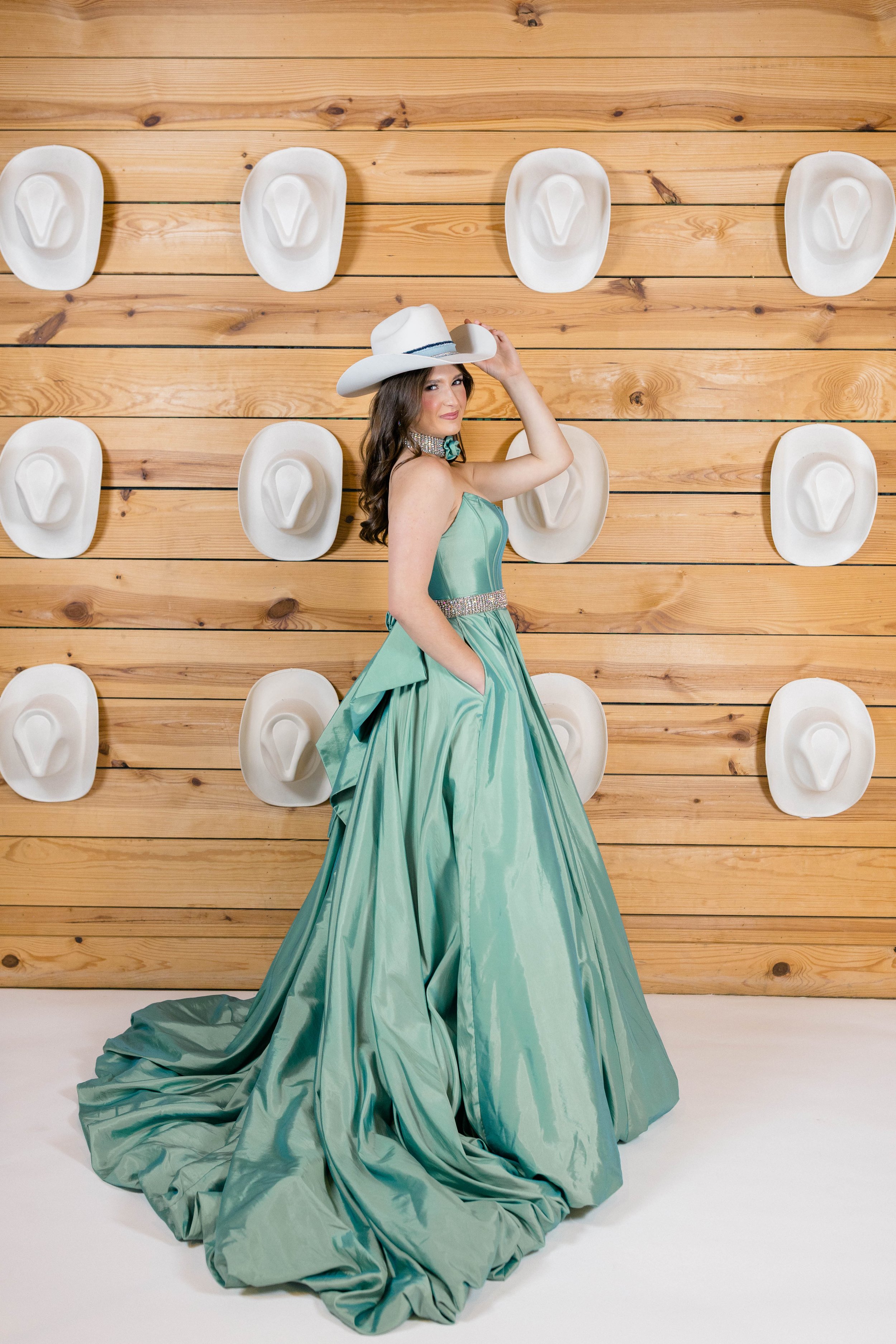 A woman in a flowing green gown with a strapless bodice and rhinestone belt, wearing a white cowboy hat with a dark band, standing in front of a wooden wall with mounted cowboy hats, posing with her hand on the hat, smiling slightly.