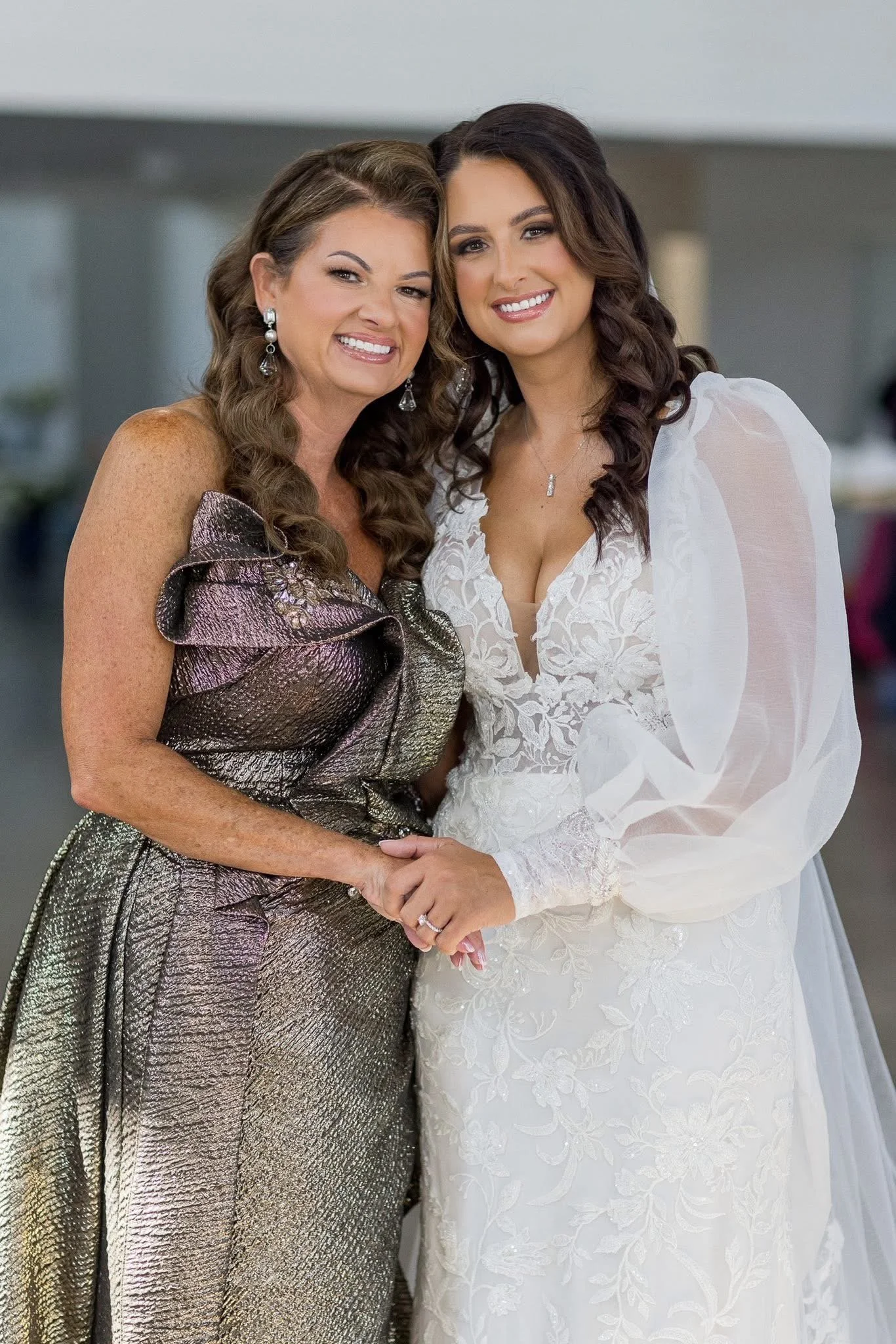 Two women, one older and one younger, smiling and holding hands at a wedding. The older woman wears a metallic dress with ruffles, and the younger woman wears a white lace wedding gown. They have long, dark hair and are in an indoor setting.