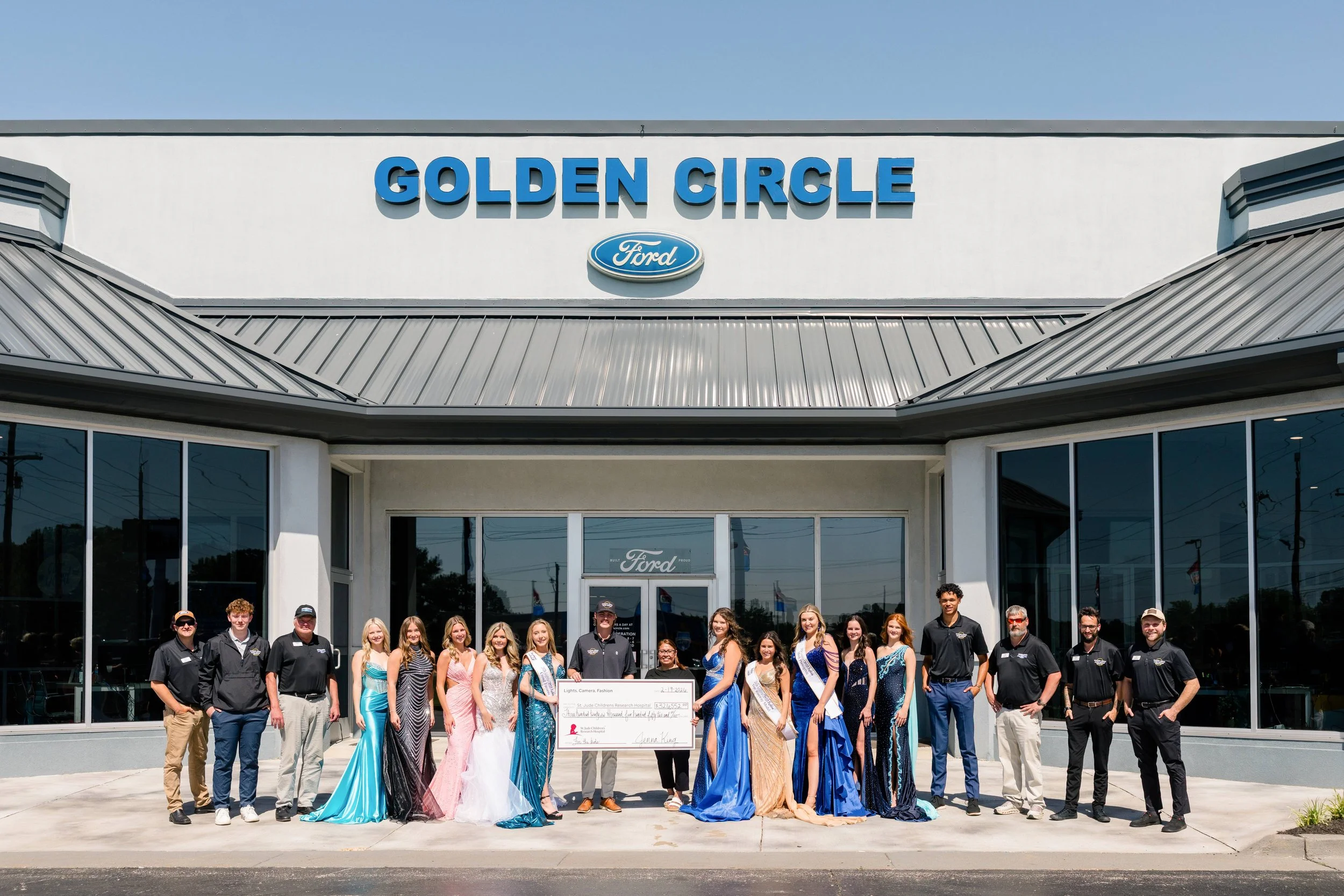 Group of young women in colorful dresses and young men, some in black uniforms, standing in front of a car dealership called Golden Circle Ford, holding a large check, with the dealership building behind them.