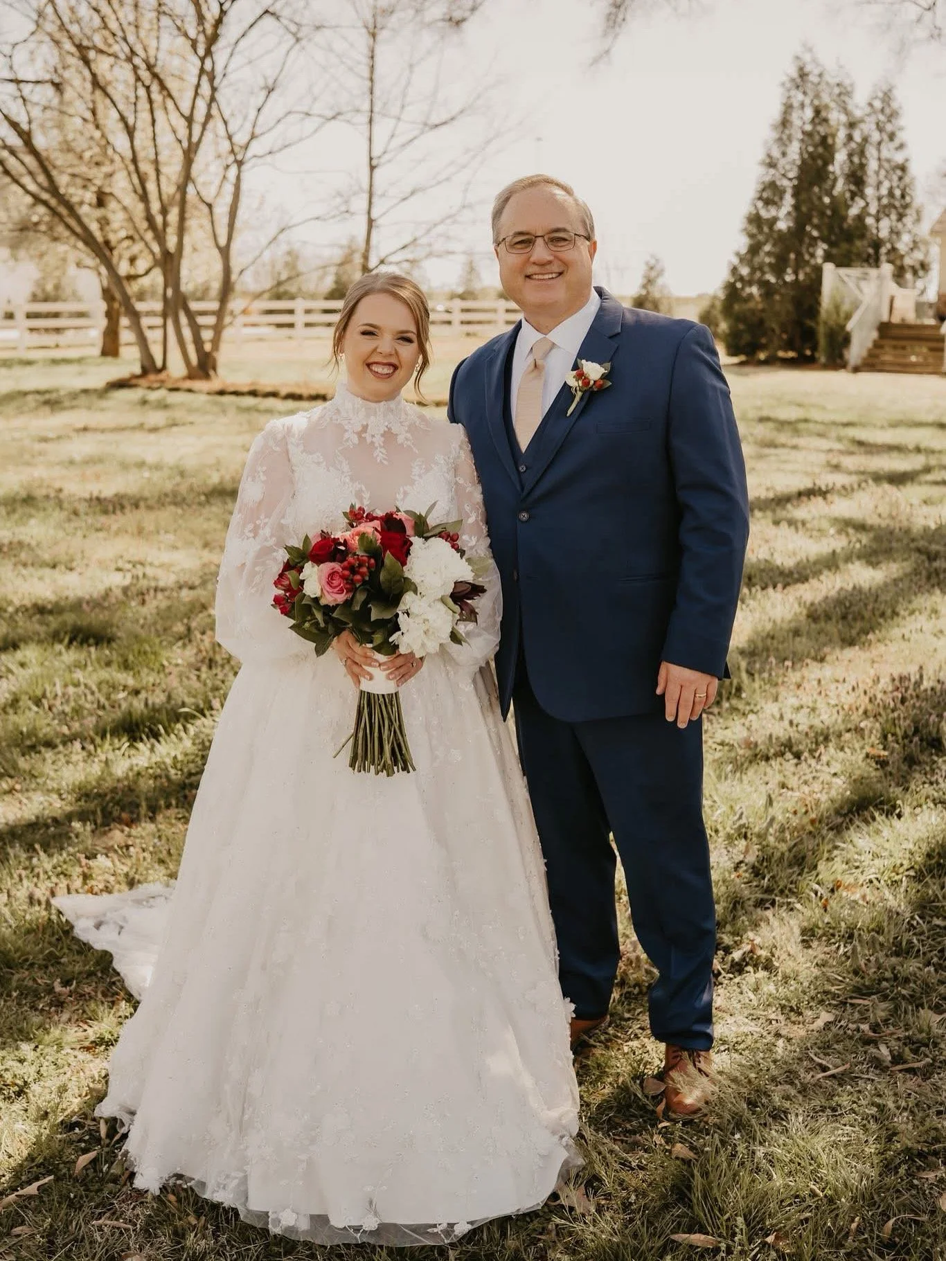 A bride in a white lace wedding dress holding a bouquet of red and white flowers, standing next to a groom in a dark blue suit outdoors during daytime.