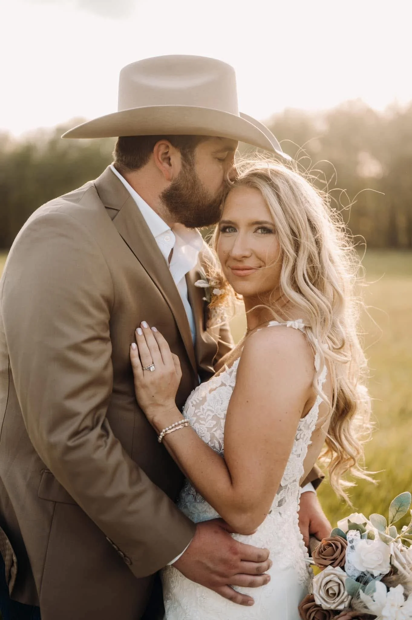 A man and woman dressed in wedding attire, with the man wearing a beige suit and wide-brimmed hat, and the woman in a white lace wedding gown, standing close outdoors in a field with blurred trees in the background. The man is kissing the woman's for