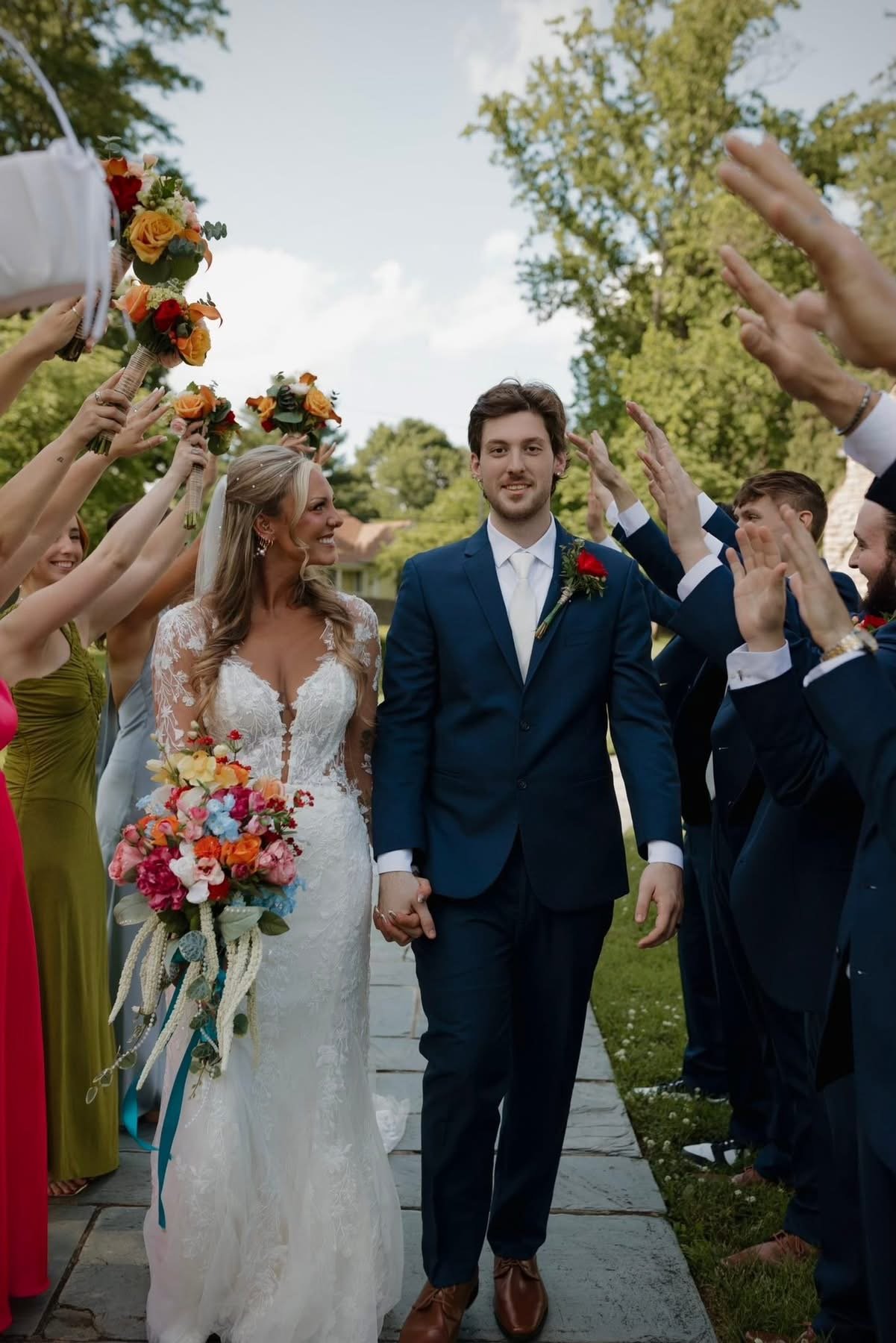 A bride and groom holding hands in a park surrounded by friends and family, celebrating a wedding, with the bride in a white lace dress and the groom in a dark blue suit, all under a blue sky with green trees.