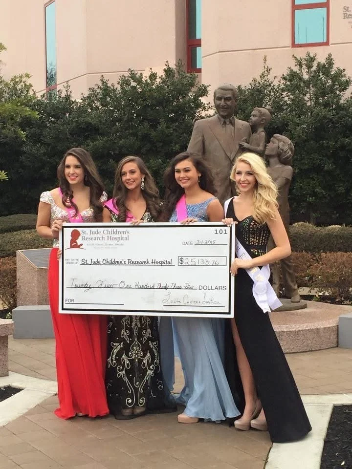 Four young women in formal dresses holding a large check for $25,133.76 made out to St. Jude Children's Research Hospital, standing outdoors in front of a statue of a man and a child, with greenery and a building in the background.